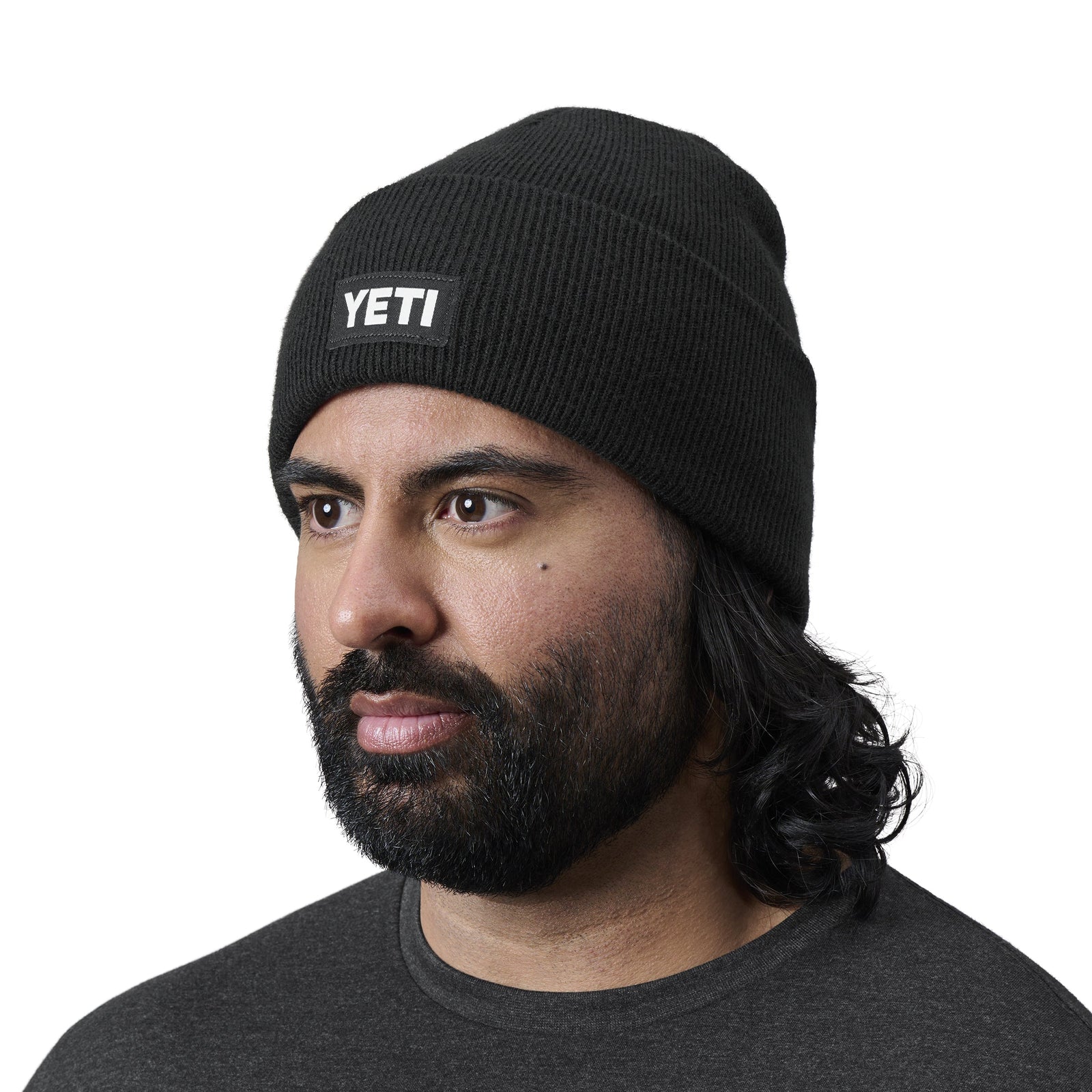 Ein Mann mit Bart und Schnurrbart trägt eine schwarze YETI® Logo Badge Beanie und blickt in einem dunklen Hemd vor einem schlichten weißen Hintergrund leicht zur Seite.