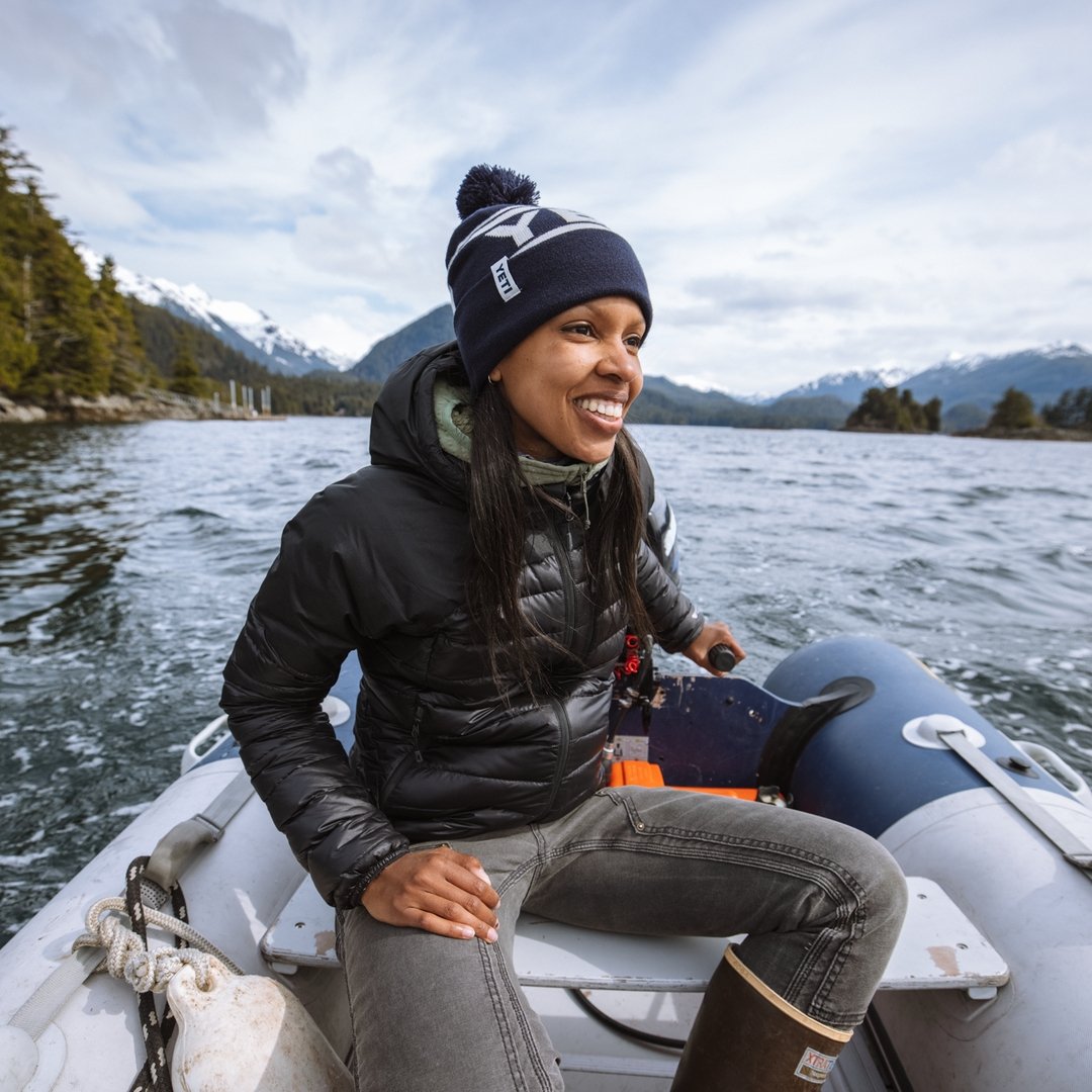 Eine lächelnde Frau mit einer marineblauen YETI® Logo Retro Knit Beanie und einer schwarzen Pufferjacke steuert ein Schlauchboot über einen Bergsee, umgeben von immergrünen und schneebedeckten Gipfeln.