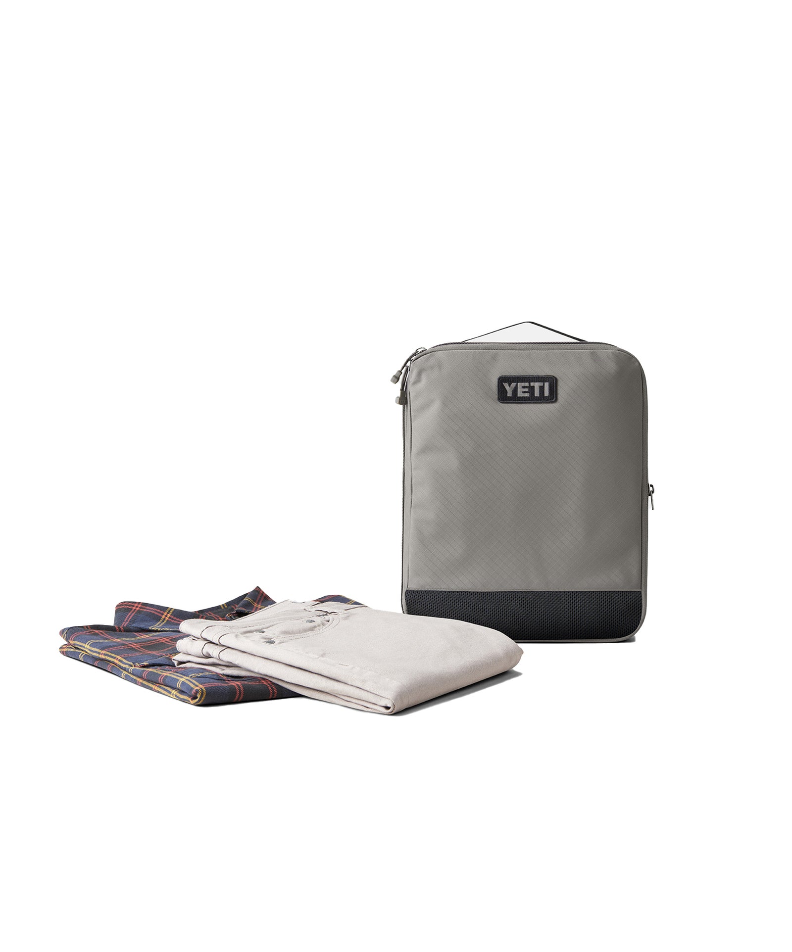 Eine YETI® Packtasche - Crossroads Cubes L in Grau steht aufrecht neben einem gefalteten karierten Hemd und einer hellen Hose auf einer weißen Fläche - perfekt als Reise Organizer für effizientes Packen.