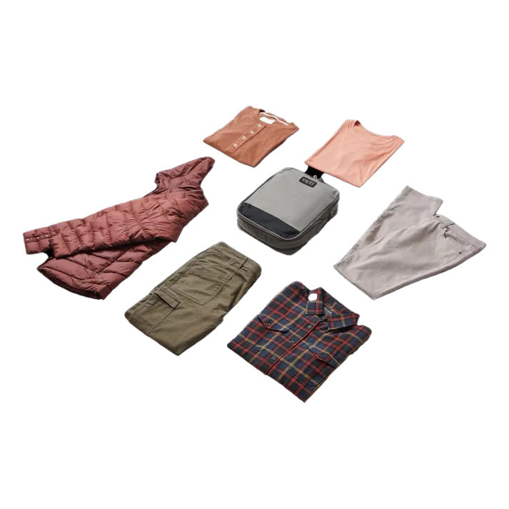 Kleidung und Reiseutensilien sind ordentlich angeordnet, darunter eine rosa Pufferjacke, zwei T-Shirts, eine graue Hose, eine grüne Hose, ein kariertes Hemd und die YETI® Packtaschen - Crossroads Cubes L - Grey von YETI auf weißem Hintergrund.