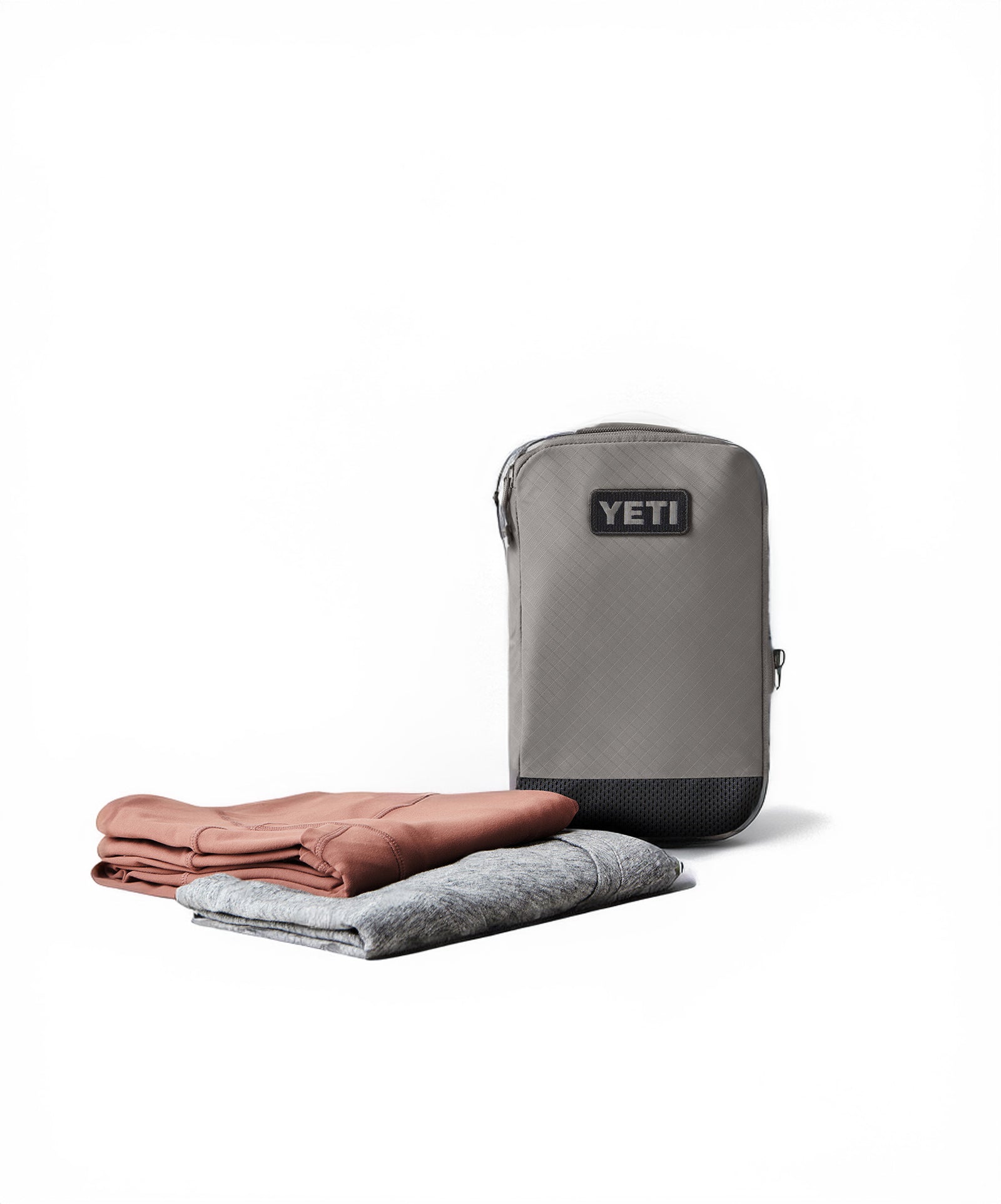Die graue YETI® Packtasche Crossroads Cubes M steht aufrecht neben zwei gefalteten Kleidungsstücken, einem pinken und einem grauen, auf weißem Hintergrund - ideal für Reisen und Organisation.
