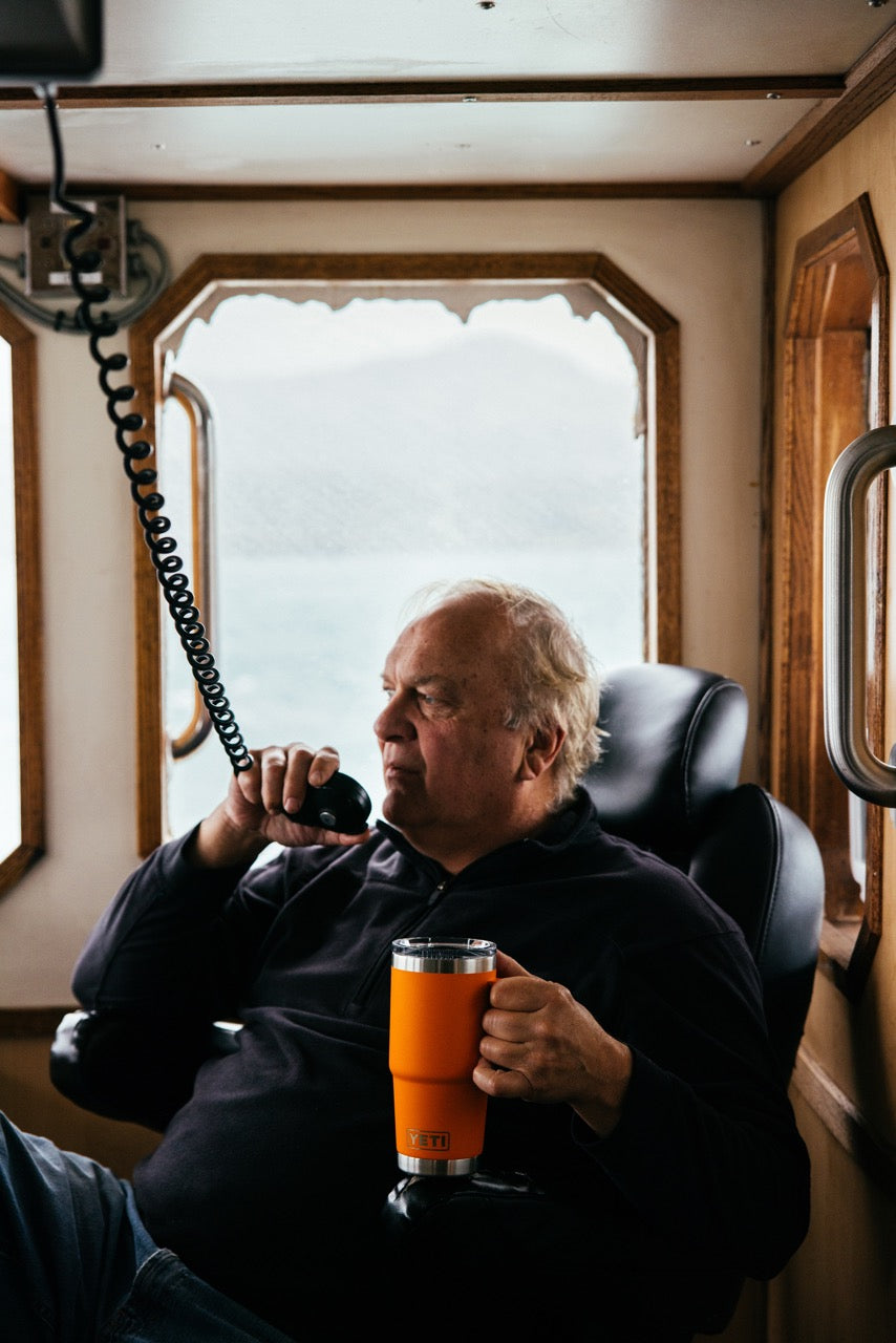 Ein älterer Mann sitzt in einer Bootskabine und hält einen YETI® Rambler 20 Oz Reisebecher - King Crab in der einen Hand, während er in ein Funkgerät spricht, während die großen Fenster hinter ihm das neblige Wasser draußen zeigen.
