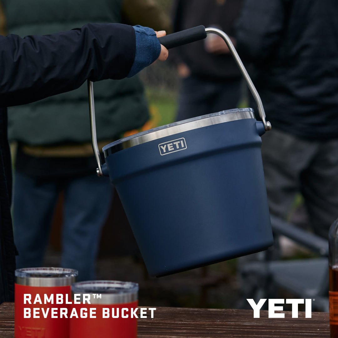 Jemand hält einen YETI® Rambler® 7,6 Liter Getränkekübel in Lowcountry Peach an seinem Metallgriff im Freien, mit roten Bechern und verschwommenen Menschen dahinter. Die doppelwandige Vakuumisolierung hält Getränke für jedes Abenteuer kalt. Text: Rambler-Getränkekübel, YETI.