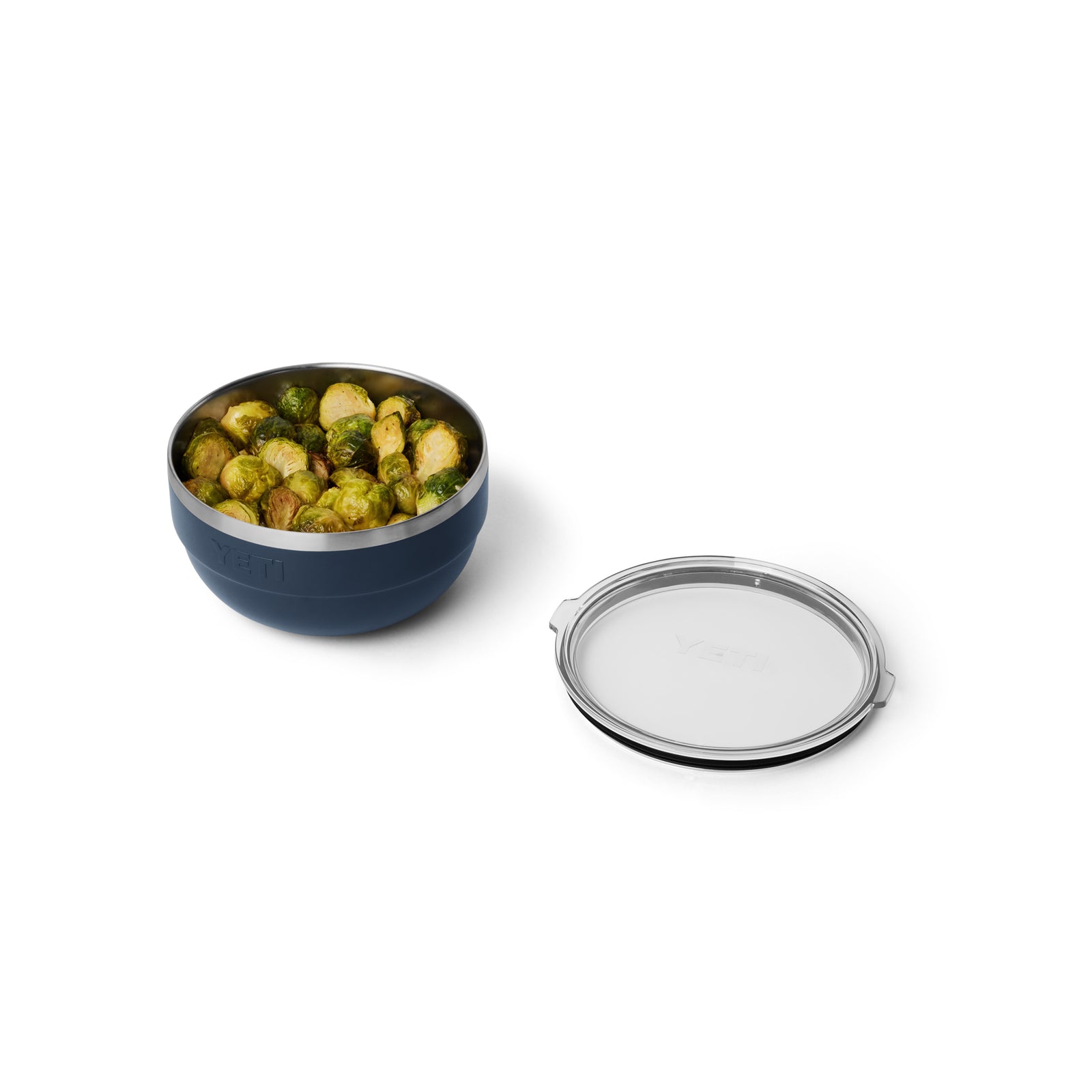 Die YETI® Rambler Bowl Medium in Navy, hergestellt von YETI aus 18/8-Edelstahl und mit doppelwandiger Vakuumisolierung, hält gerösteten Rosenkohl neben dem durchsichtigen Deckel auf weißem Hintergrund.