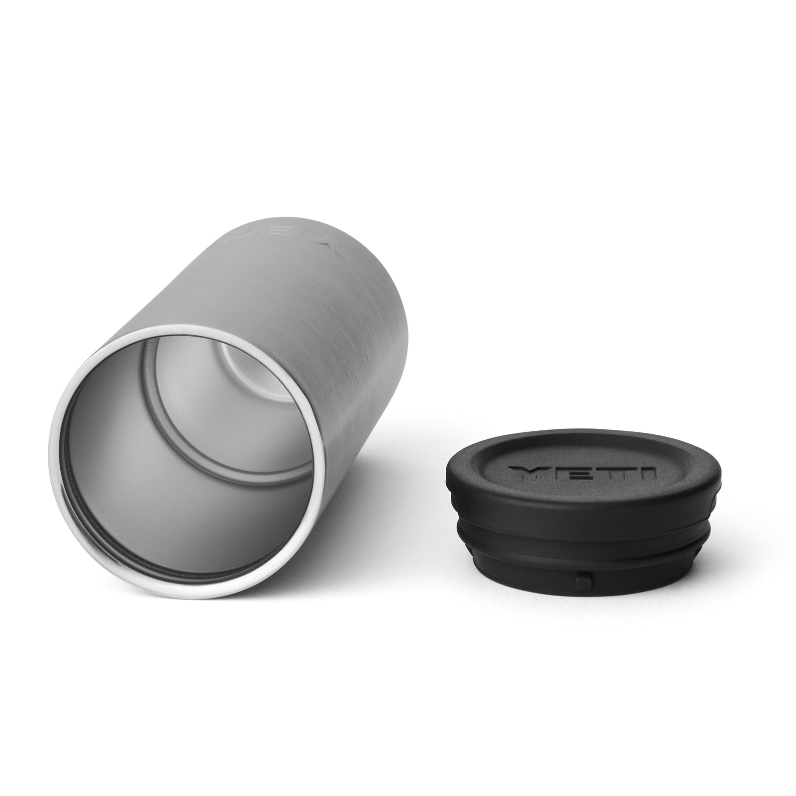 Ein YETI® Rambler Schnapsbecher aus Edelstahl liegt seitlich neben einem schwarzen, runden Deckel mit geprägtem YETI-Logo. Das offene Ende zeigt das glatte Edelstahl-Innenleben des Bechers.