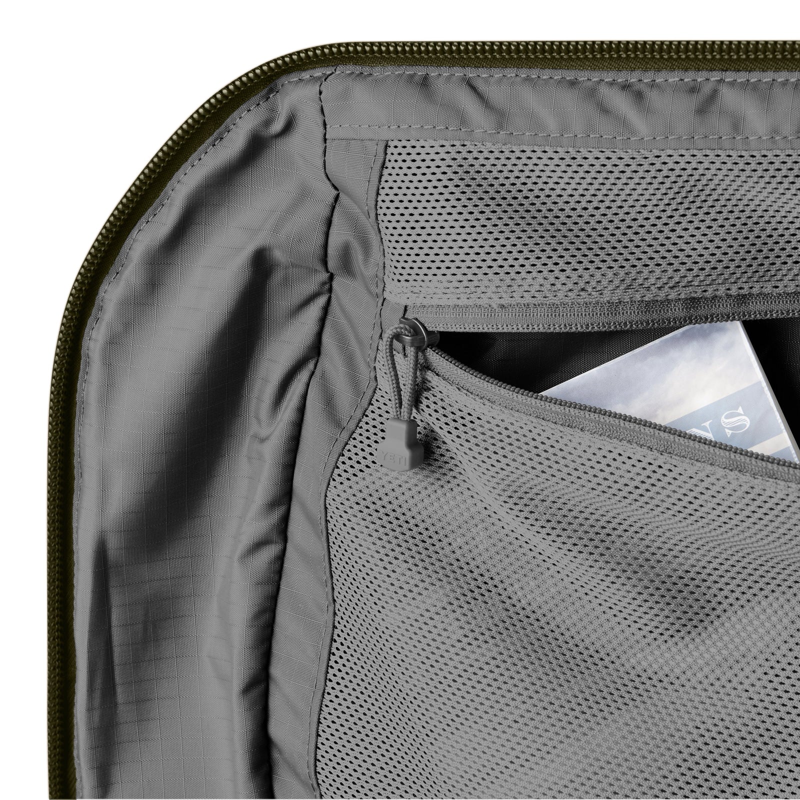 Nahaufnahme des Innenraums der olivgrünen YETI® Reisetasche Crossroads® 40L mit einer Netztasche mit Reißverschluss, in der sich ein Magazin befindet. Das TuffSkin-Nylon-Material sieht robust aus und unterstreicht das saubere und organisierte Design der Tasche.