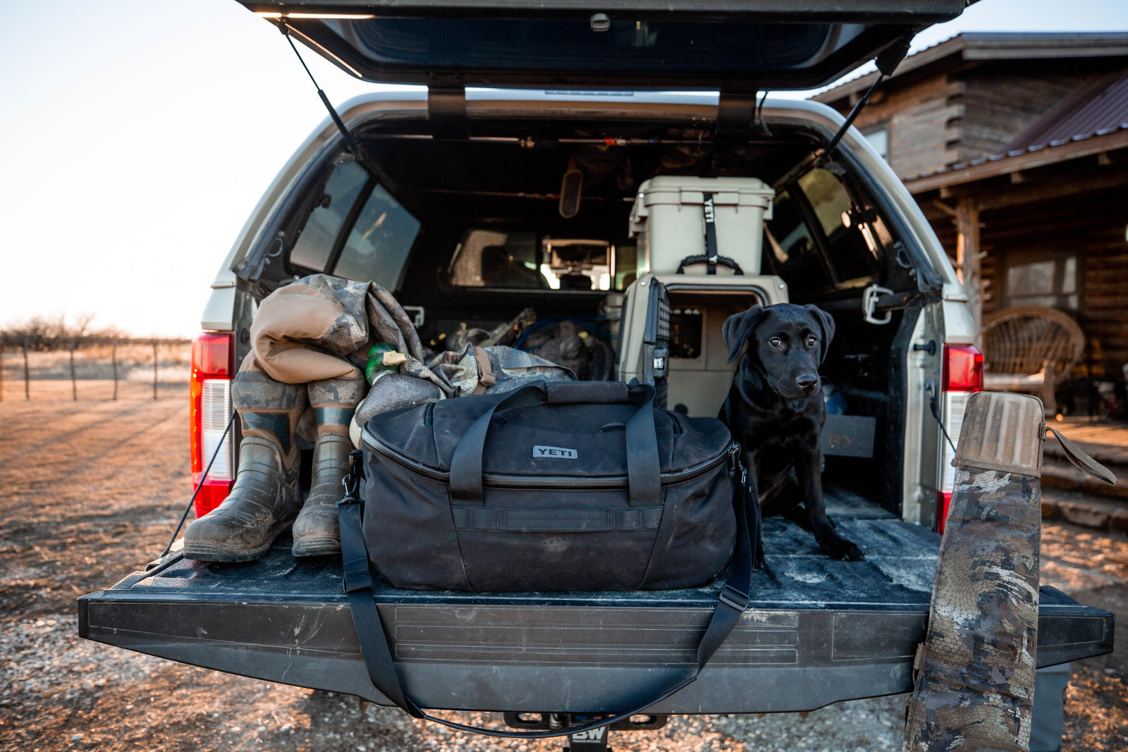 Ein schwarzer Hund sitzt auf der offenen Heckklappe eines Lastwagens neben der YETI® Reisetasche Crossroads 60 L - Black, schlammigen Stiefeln und Outdoor-Ausrüstung, mit einem rustikalen Haus und trockener Landschaft im Hintergrund.