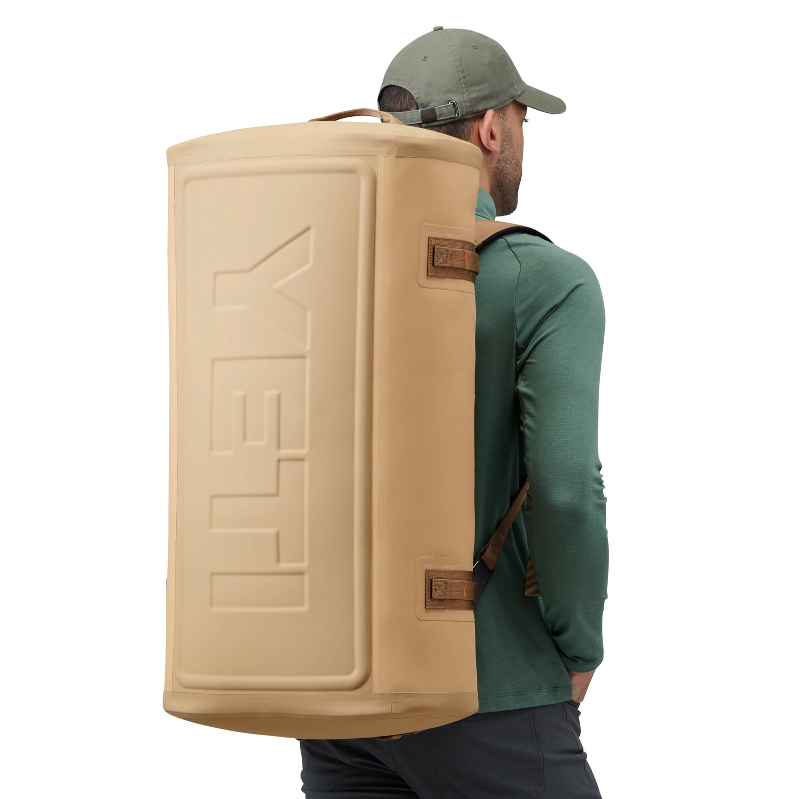 Ein Mann mit grüner Mütze und Hemd trägt die YETI® Reisetasche Panga 75 - Tan mit Hilfe von Schulterriemen auf dem Rücken. Die Tasche, die sich perfekt für Outdoor-Abenteuer eignet, trägt das kultige YETI-Logo auf der Seite.