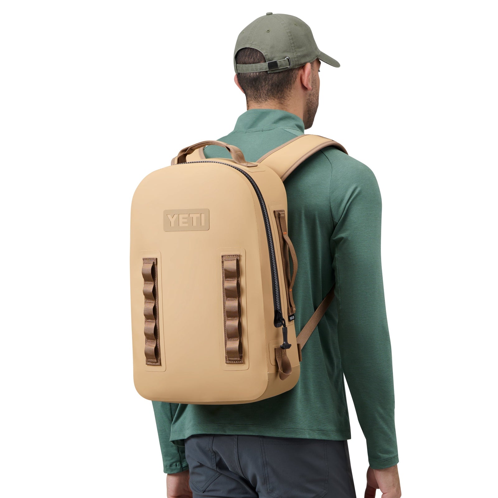 Eine Person, die ein grünes langärmeliges Hemd, eine graue Hose und eine grüne Mütze trägt, ist von hinten mit dem YETI® Rucksack Panga 28 - Tan von YETI vor einem schlichten weißen Hintergrund zu sehen.