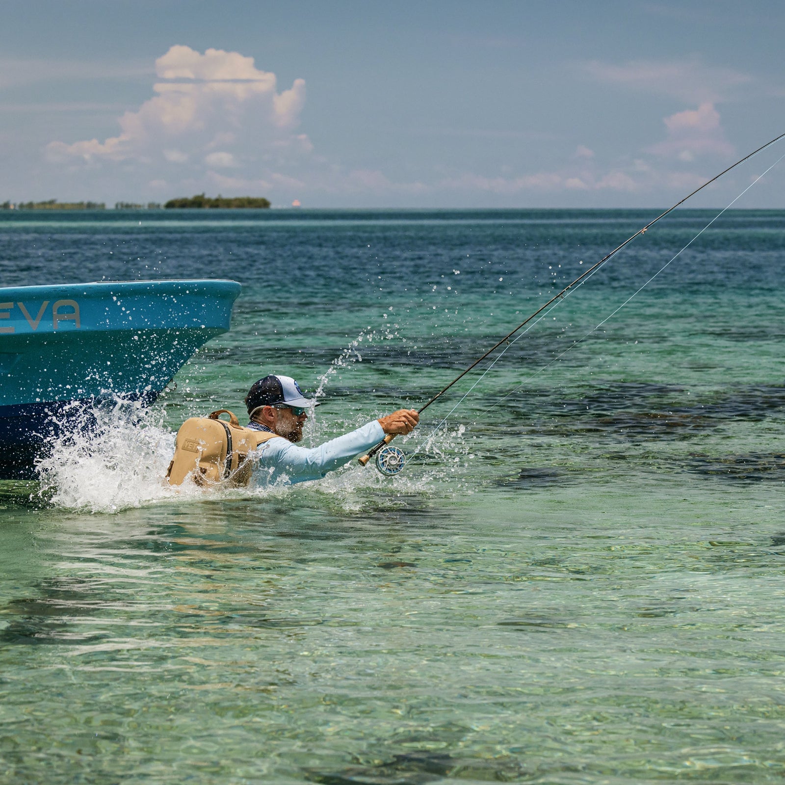 Eine Person mit Mütze und YETI® Rucksack Panga 28 - Tan watet beim Fliegenfischen durch klares, seichtes Wasser in der Nähe eines blauen Bootes und plätschert, während sich der Ozean unter teilweise bewölktem Himmel bis zu den entfernten Inseln erstreckt.