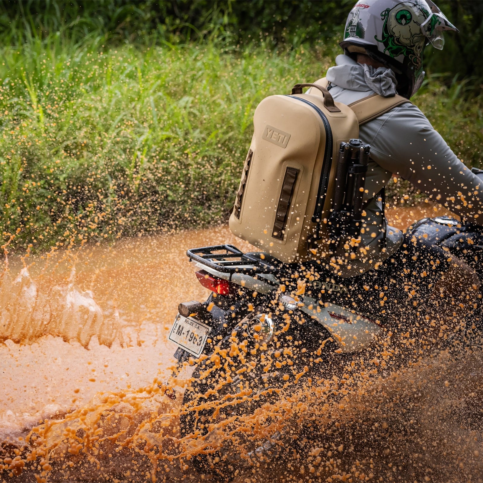 Ein behelmter Fahrer mit einem YETI® Rucksack Panga 28 - Tan rast durch eine schlammige Pfütze und lässt orange-braune Gischt aufsteigen. Der wasserdichte YETI-Rucksack ist für Abenteuer inmitten von saftig grünem Gras im Hintergrund gebaut.