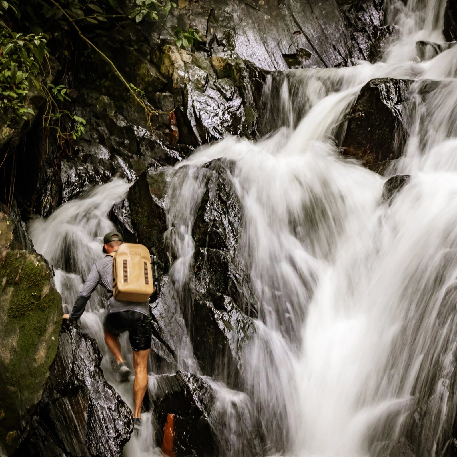 Eine Person mit einem braunen YETI® Rucksack Panga 28 klettert auf nasse, dunkle Felsen neben einem rauschenden Wasserfall in einem üppigen, grünen Wald.