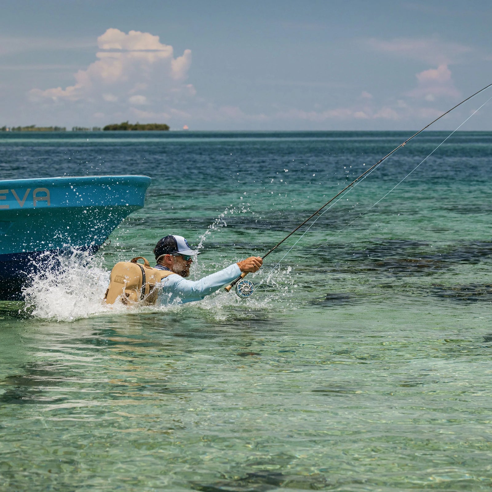 Ausgestattet mit Outdoor-Ausrüstung und dem YETI® Rucksack Panga 28 - Tan steht ein Angler im flachen, klaren Wasser neben einem blauen Boot und wirft seine Angel aus, während das Wasser um ihn herum unter einem sonnigen Himmel plätschert.
