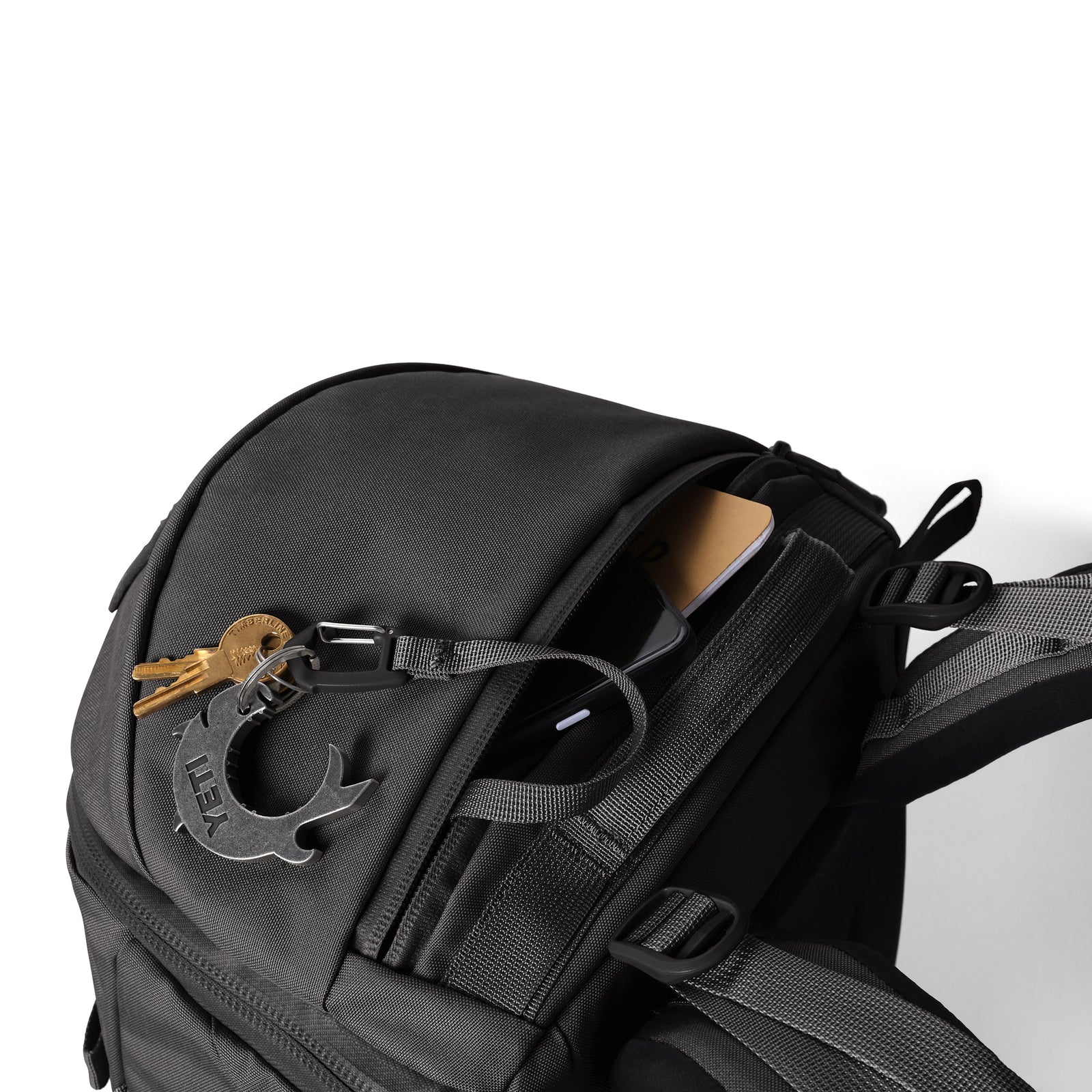 Ein schwarzer YETI® Rucksack Ranchero™ 22L steht vor einem weißen Hintergrund, der teilweise geöffnet wurde, um ein Telefon und ein Notizbuch in der oberen Tasche zu enthüllen. Am Reißverschluss dieses wasserdichten Rucksacks von YETI hängen Schlüssel mit einem Mondsichelanhänger.