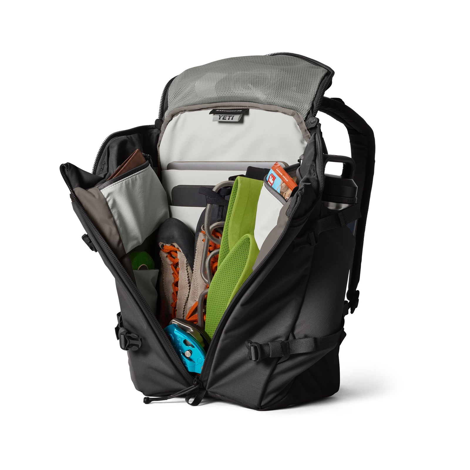 Ein schwarzer YETI® Rucksack Ranchero™ 22L wird aufgeklappt gezeigt, mit geordneten Fächern, gefüllt mit Kletterschuhen, einem Klettergurt, einer Wasserflasche, einem Helm und diverser Outdoor-Ausrüstung.