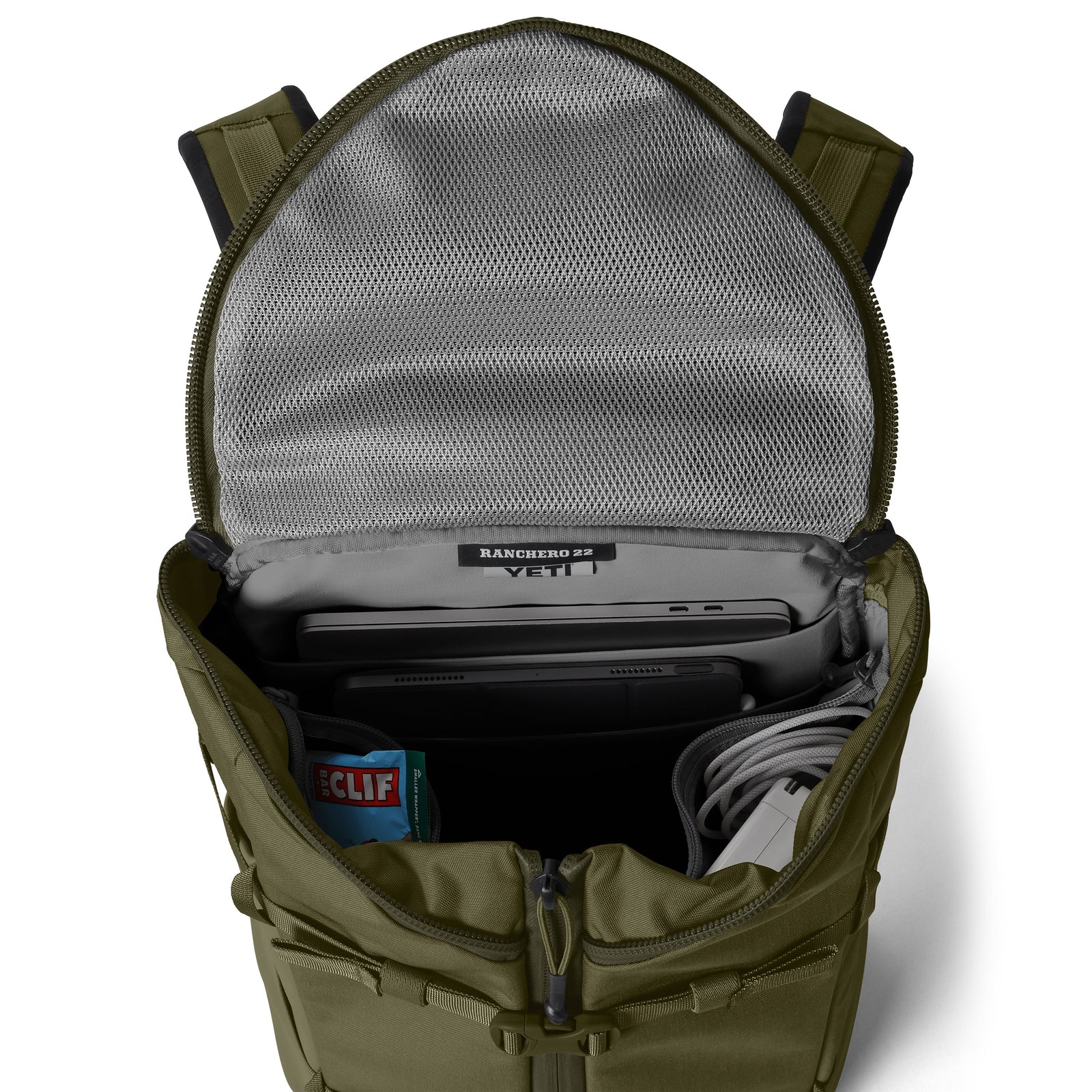 Ein geöffneter YETI® Rucksack Ranchero™ 22L - Olive zeigt einen Laptop, ein Tablet, Kabel und eine CLIF-Bar im Inneren. Er ist aus TuffSkin-Nylon gefertigt und bietet Netztaschen, organisierte Fächer und ein Laptop-Fach zur sicheren Aufbewahrung.