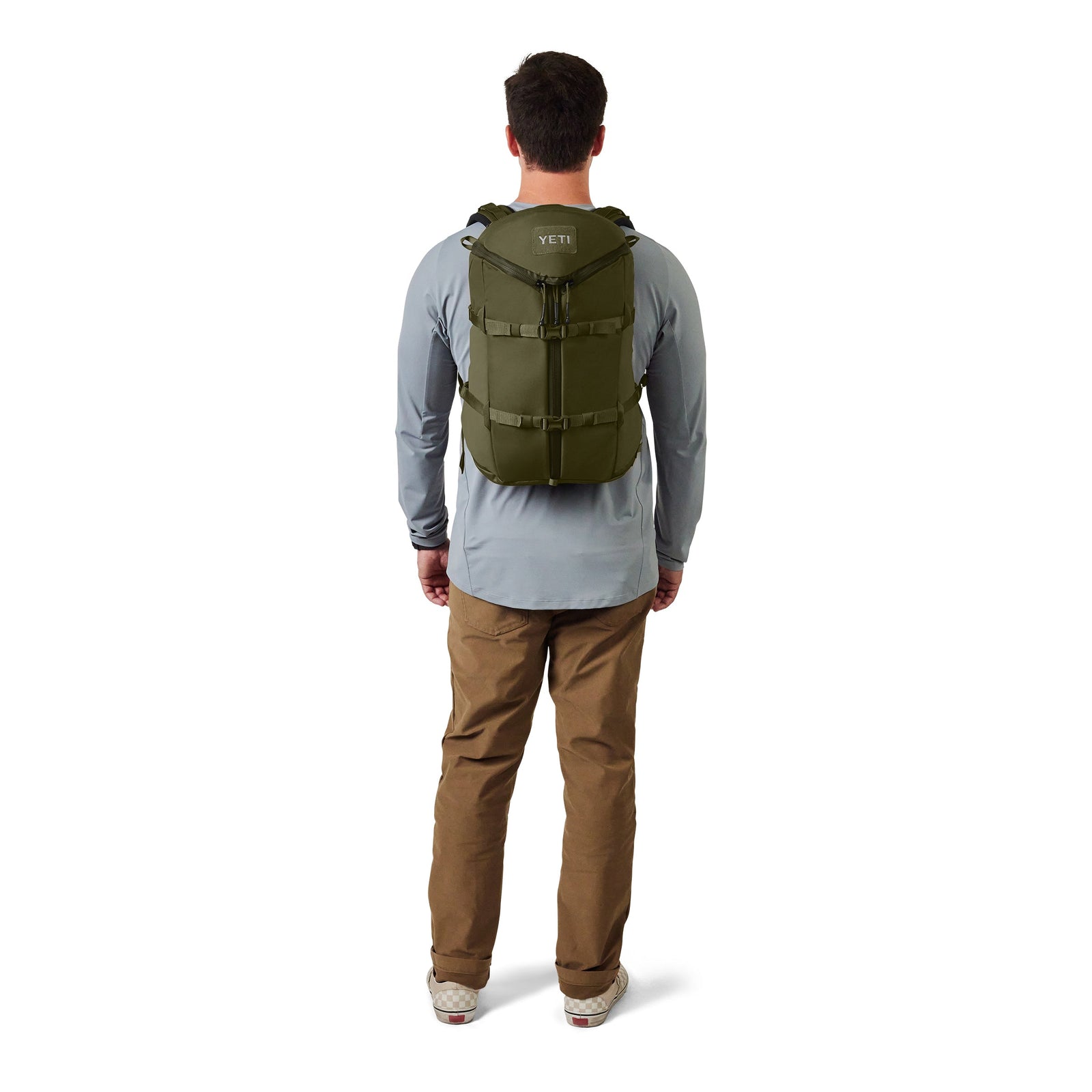 Eine Person steht mit einem olivgrünen YETI® Rucksack Ranchero™ 22L aus TuffSkin-Nylon, einem hellgrauen langärmeligen Hemd, einer khakifarbenen Hose und weißen Turnschuhen vor einem einfarbig weißen Hintergrund.