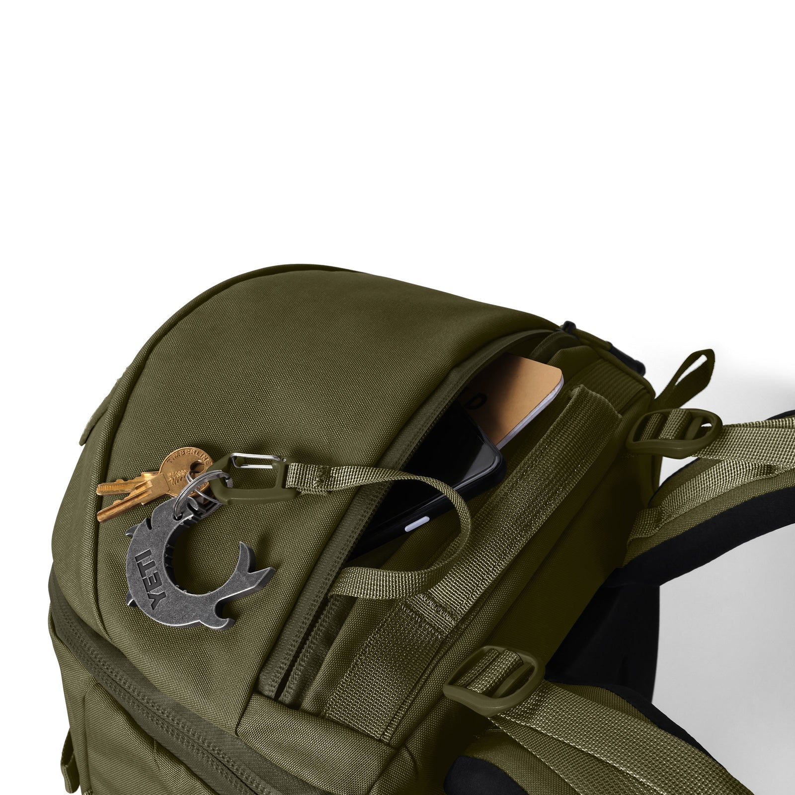Der olivgrüne YETI® Rucksack Ranchero™ 22L ist teilweise geöffnet und zeigt ein Telefon und eine Brieftasche im Inneren. Am Reißverschluss hängen Schlüssel und ein Multitool aus Metall, während ein spezielles Laptopfach Ihre Technik schützt.