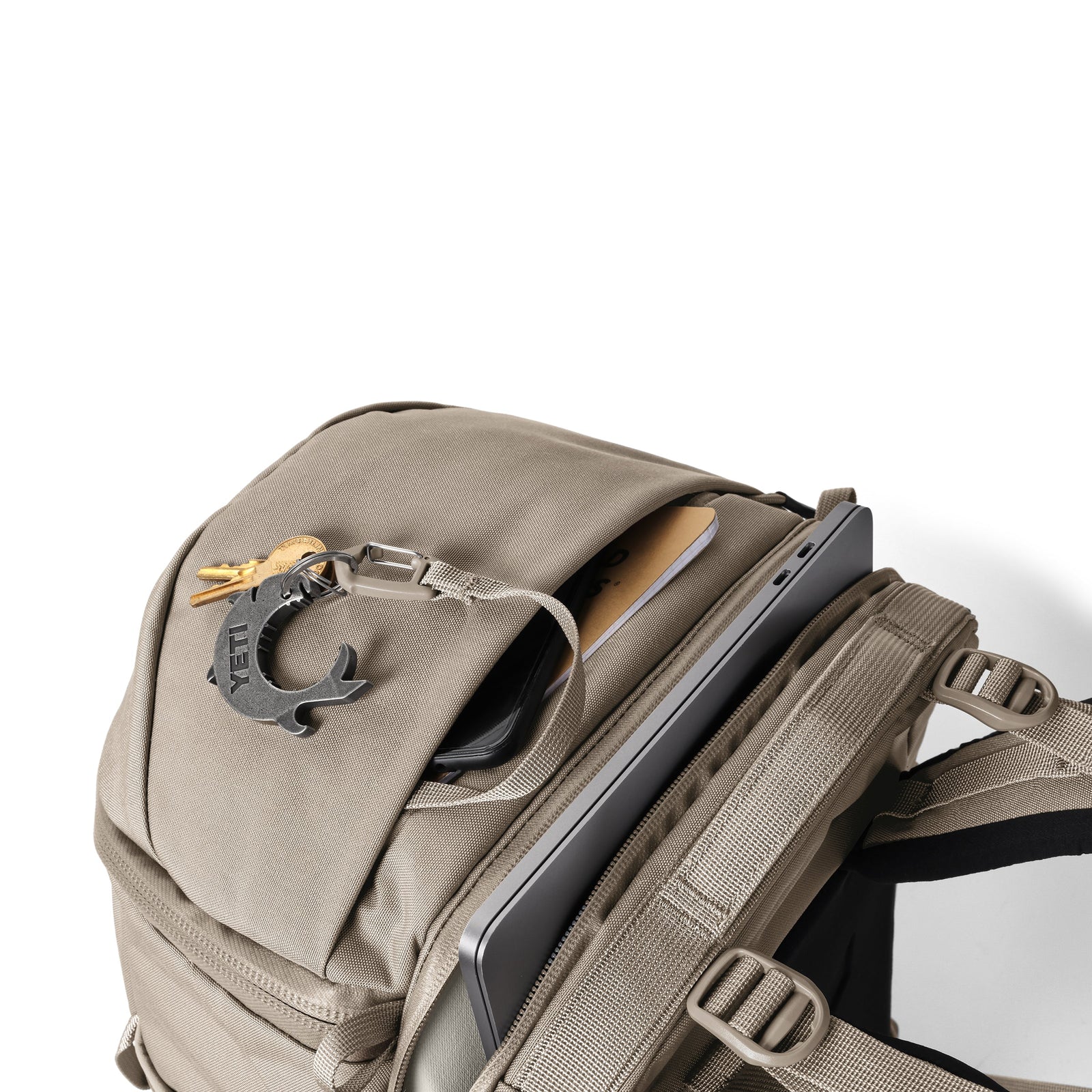 Ein YETI® Rucksack Ranchero™ 27L in Cape Dark Taupe mit einem Laptop und einem Tablet in separaten Fächern. Ein schwarzer Schlüsselbund und Schlüssel hängen am oberen Reißverschluss. Diese wasserfeste YETI-Tasche ist vor einem weißen Hintergrund abgebildet.