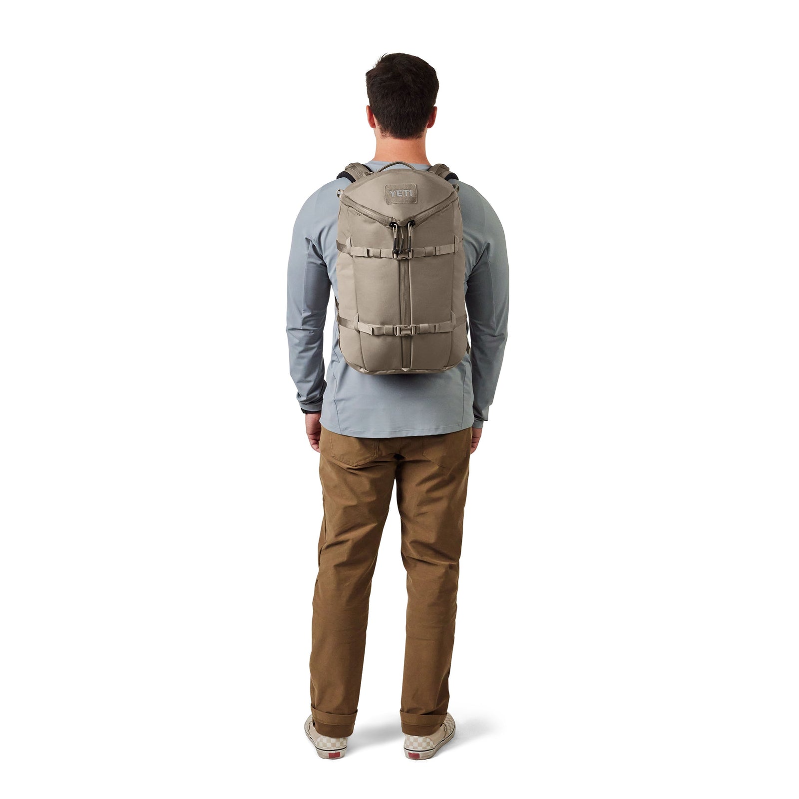 Eine Person mit einem YETI® Rucksack Ranchero™ 27L - Cape Dark Taupe, einem hellblauen langärmeligen Hemd, einer khakifarbenen Hose und Turnschuhen steht vor einem weißen Hintergrund.