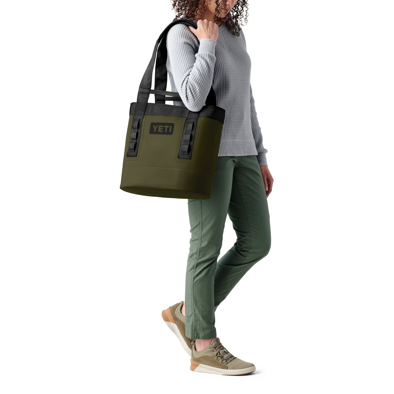 Eine Person in grauem Pullover, grüner Hose und hellbraunen Turnschuhen trägt eine große olivgrüne YETI® Tragetasche Camino® Carryall 20 - eine elegante wasserdichte Tasche mit schwarzen Griffen - auf der Schulter.