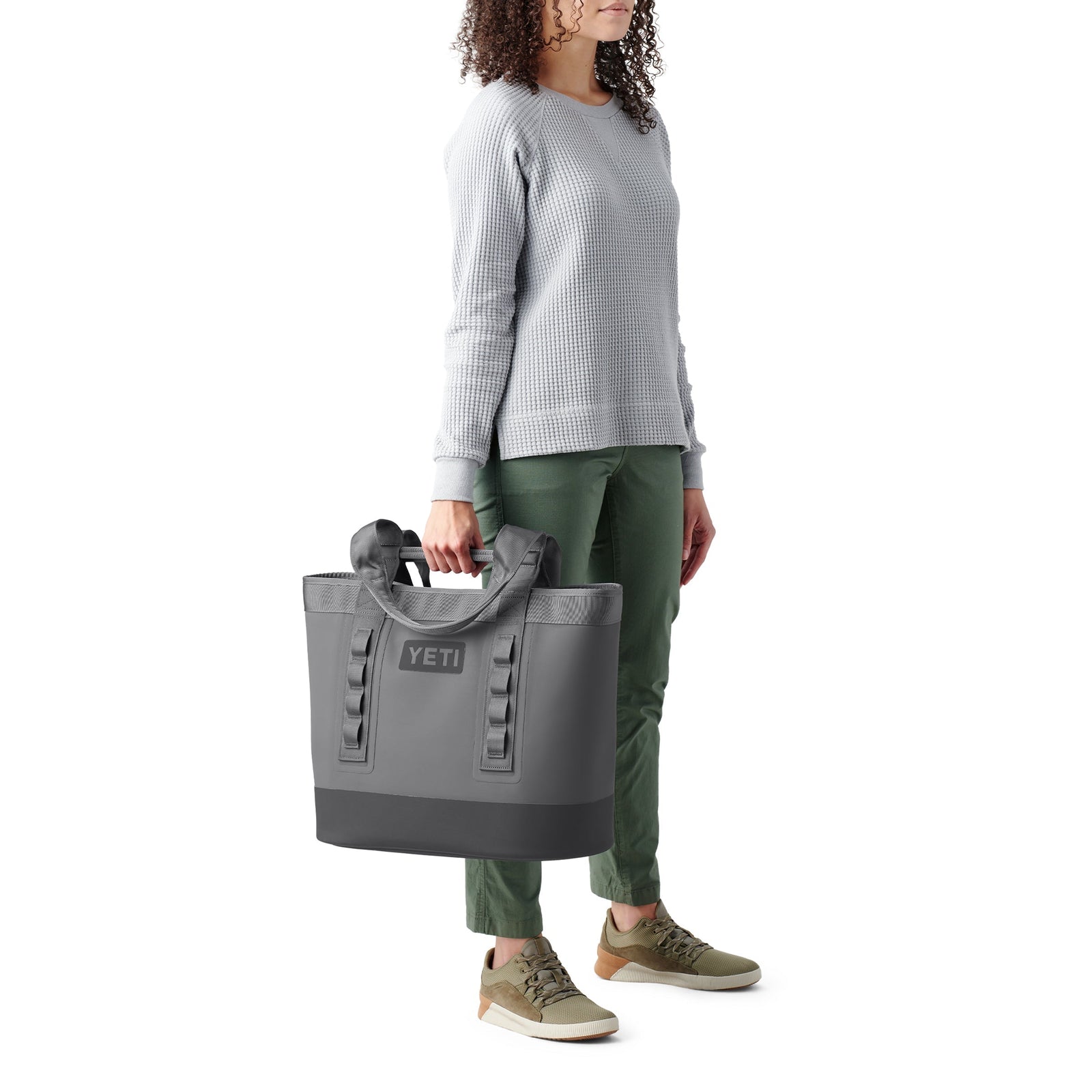 Eine Person in einem hellgrauen Pullover, einer grünen Hose und olivgrünen Turnschuhen hält die YETI® Tragetasche Camino® Carryall 35 - Navy vor einem weißen Hintergrund an ihrer Seite.