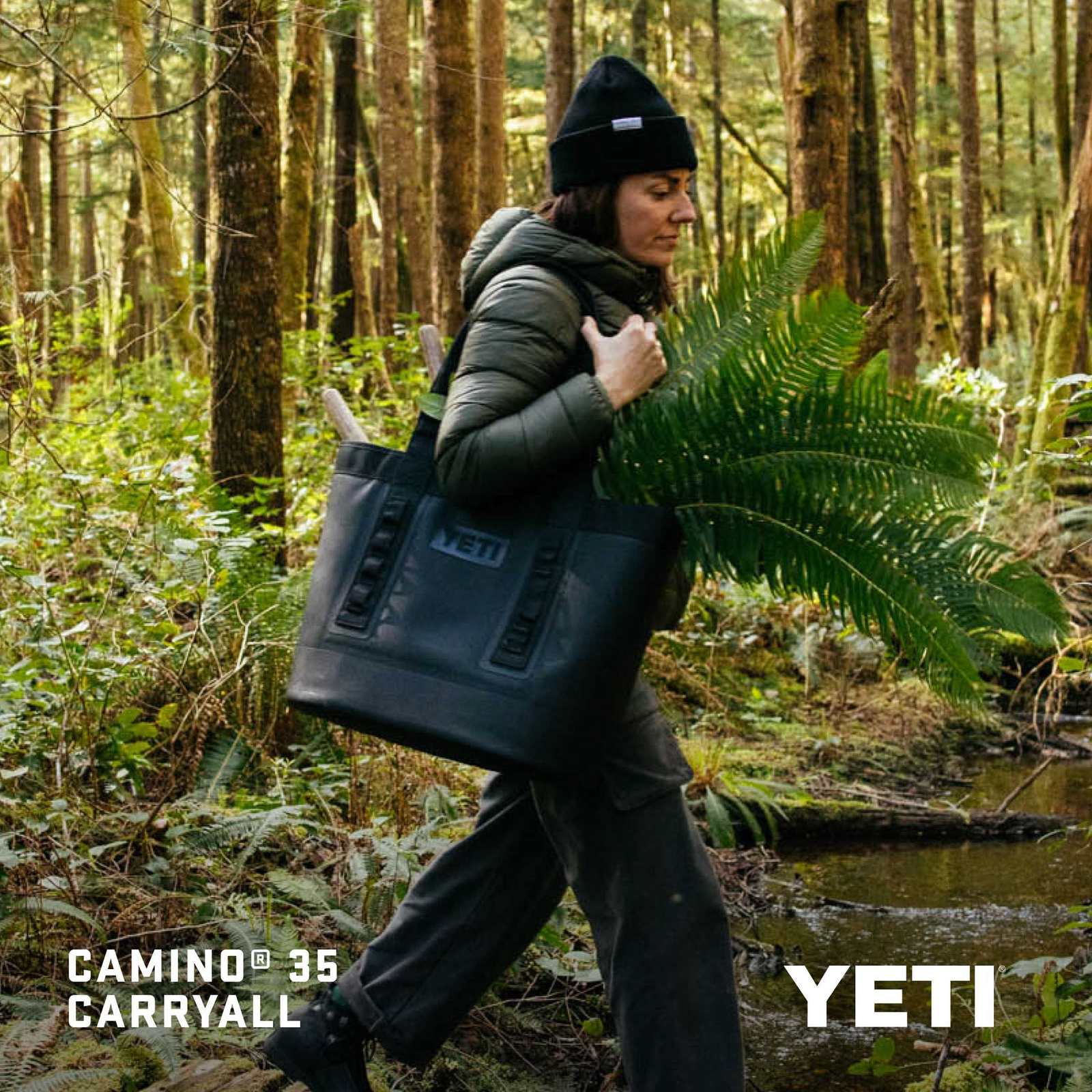 Eine Person in Outdoor-Ausrüstung trägt eine YETI® Tragetasche Camino® Carryall 35 - Navy, gefüllt mit Farnen und Werkzeug, durch einen Wald. Das Bild zeigt "CAMINO® 35 CARRYALL" und "YETI" in fetter weißer Schrift.