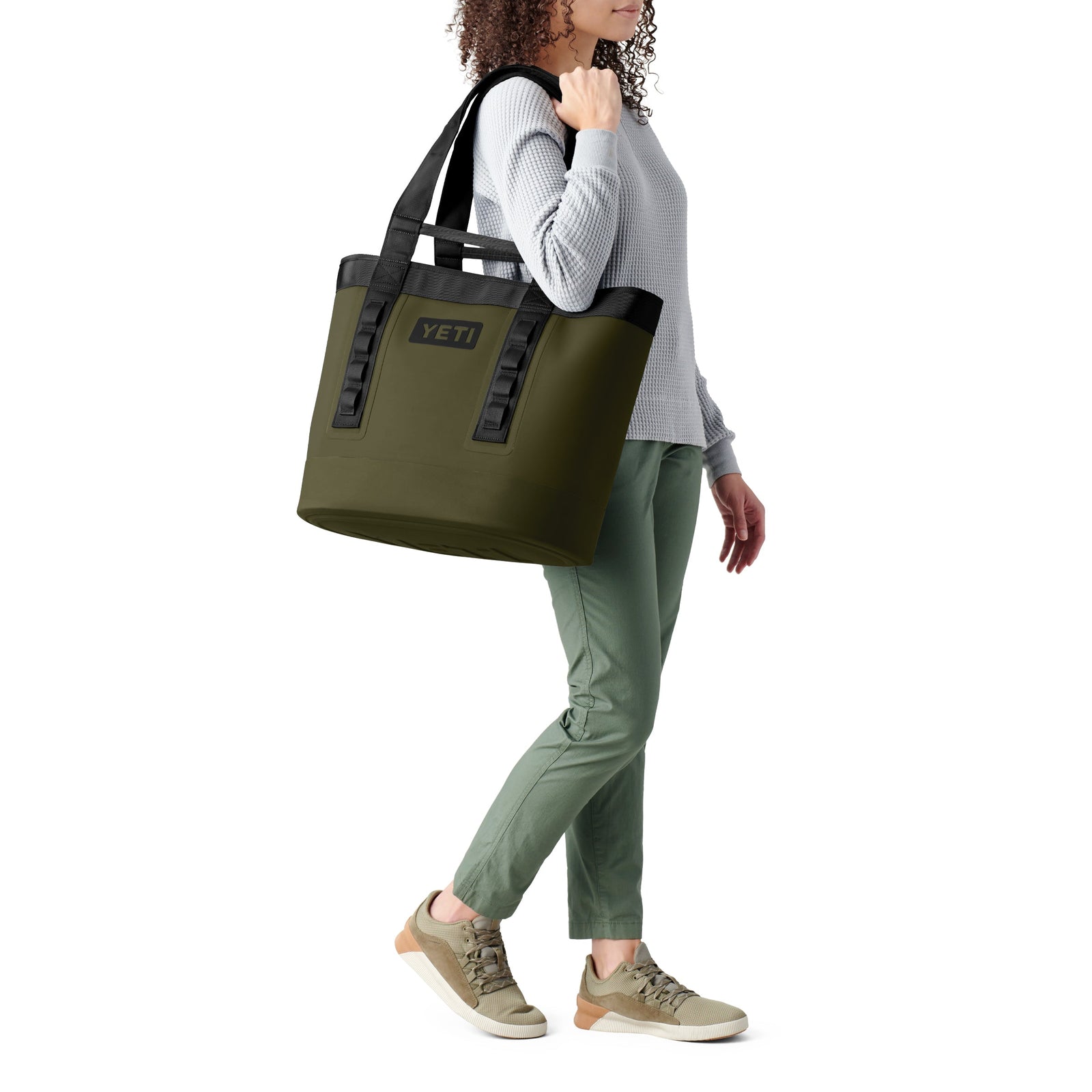 Eine Person steht vor einem schlichten weißen Hintergrund und trägt die YETI® Tragetasche Camino® Carryall 35 - Olive über der Schulter. Sie trägt einen grauen strukturierten Pullover, eine grüne Hose und beigefarbene Turnschuhe.