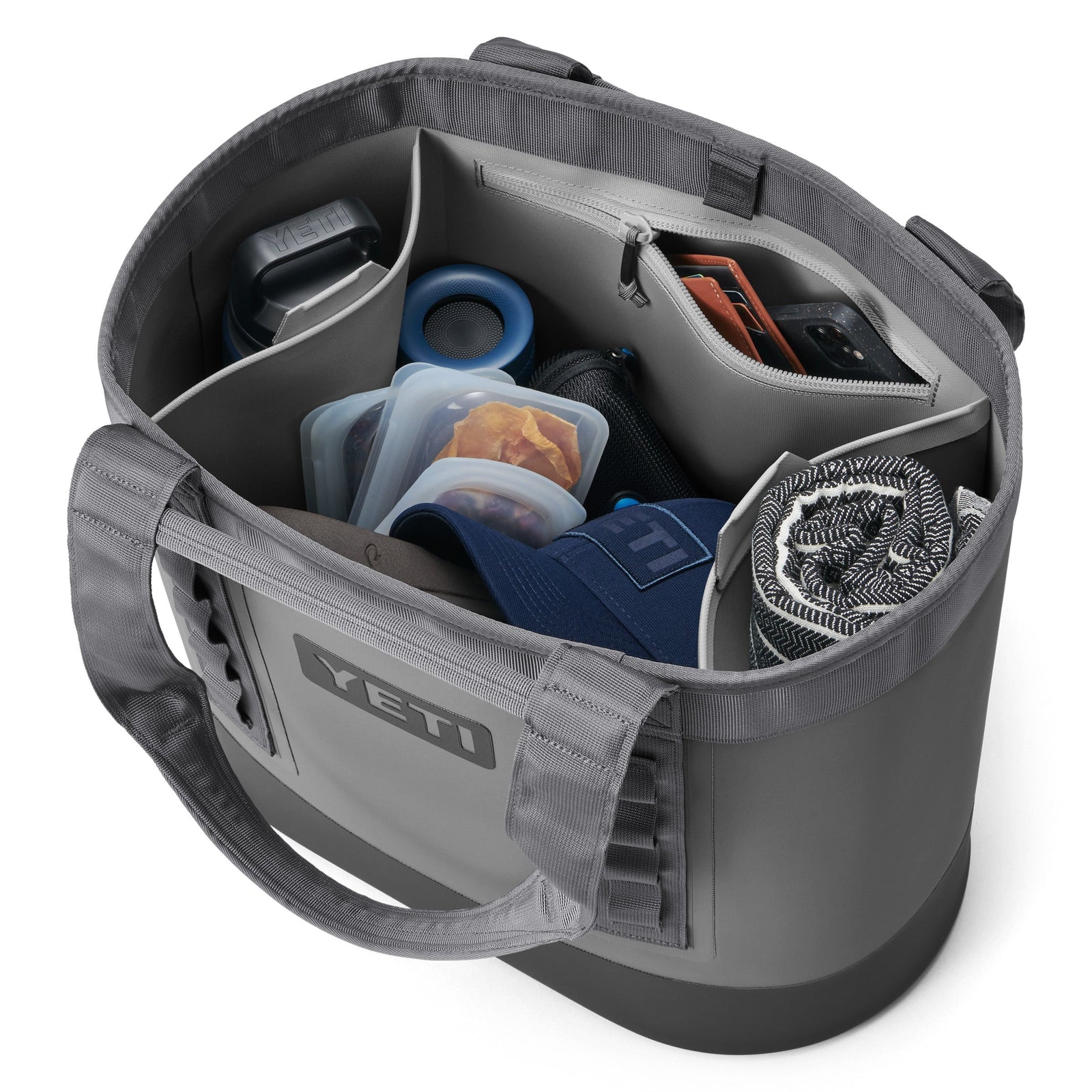 Eine YETI® Tragetasche Camino® Carryall 35 in Storm Grey, mit mehreren Fächern, zeigt sichtbar Wasserflasche, Mütze, Essensbehälter, zusammengerolltes Handtuch, Sonnenbrille und Handy - alles ordentlich organisiert und griffbereit.