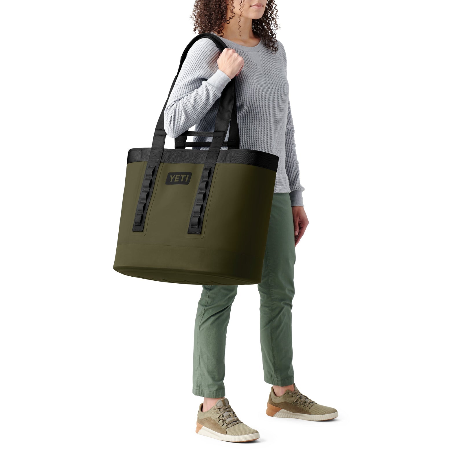 Eine Person in einem hellgrauen Pullover, einer grünen Hose und hellbraunen Turnschuhen trägt die YETI® Tragetasche Camino® Carryall 50 - Olive über der Schulter vor einem weißen Hintergrund.