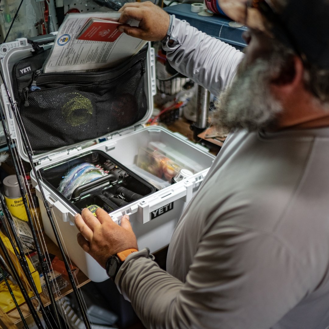 Ein bärtiger Mann mit Sonnenbrille öffnet eine YETI® LoadOut GoBox 30 mit der YETI® Trennwand und hält ein rotes Handbuch über die Angelausrüstung und die Köder, die in seiner Werkstatt fein säuberlich angeordnet sind.