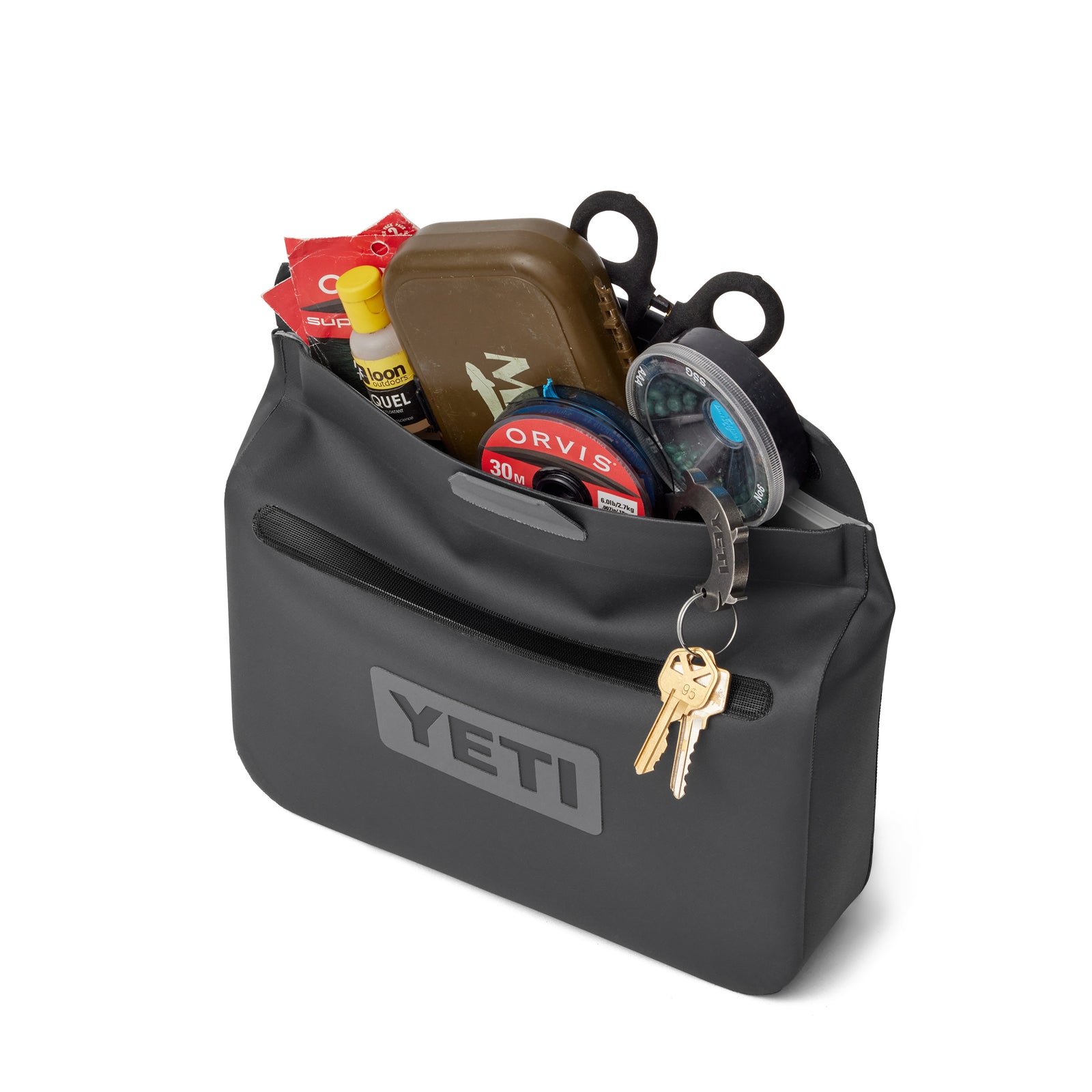 Eine YETI® Zubehörtasche - Sidekick® 3L in Charcoal, gefüllt mit Angelzubehör wie Schere, Angelschnur, braunem Etui und Dosen. Zwei Schlüssel am Ring sind vorne am Reißverschluss befestigt. Perfekt als Trekking-Zubehör.