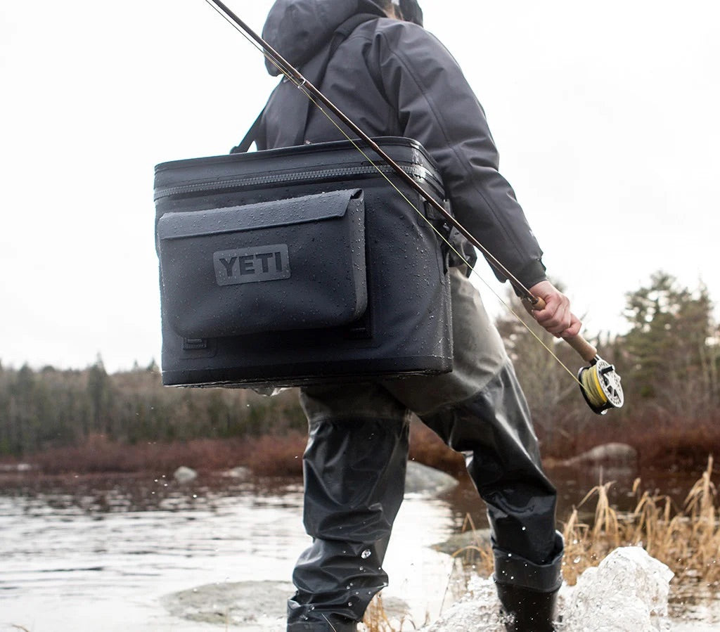 Eine Person in Wathose steht im flachen Wasser eines Sees und hält eine Angelrute mit der YETI® Zubehörtasche - Sidekick® 3L - Charcoal über der Schulter. Im Hintergrund sind Bäume und ein bewölkter Himmel zu sehen.