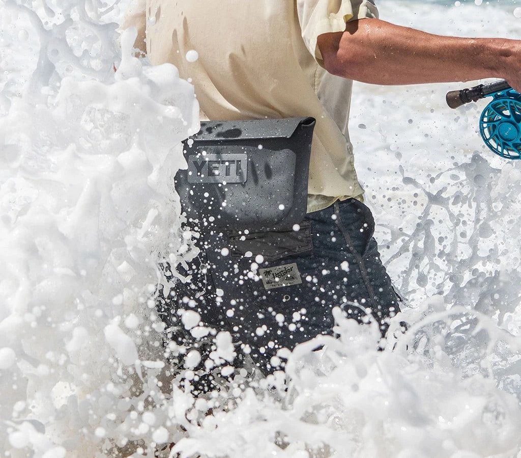 Eine Person steht in den plätschernden Wellen des Ozeans, hält eine blaue Angel in der Hand und trägt ein beiges Hemd mit einer YETI® Zubehörtasche - Sidekick® 3L - Charcoal von YETI um die Hüfte.