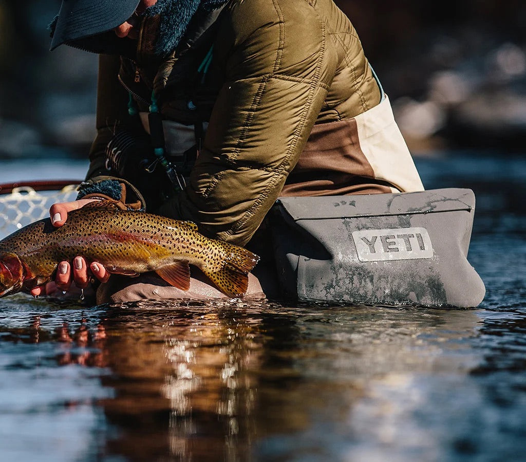 Eine Person kniet im flachen Wasser und hält einen bunten Fisch, während sie eine Outdoor-Ausrüstung und eine Jacke trägt. Auf ihrer Hüfte ruht eine YETI® Zubehörtasche - Sidekick® 3L in Charcoal von YETI.