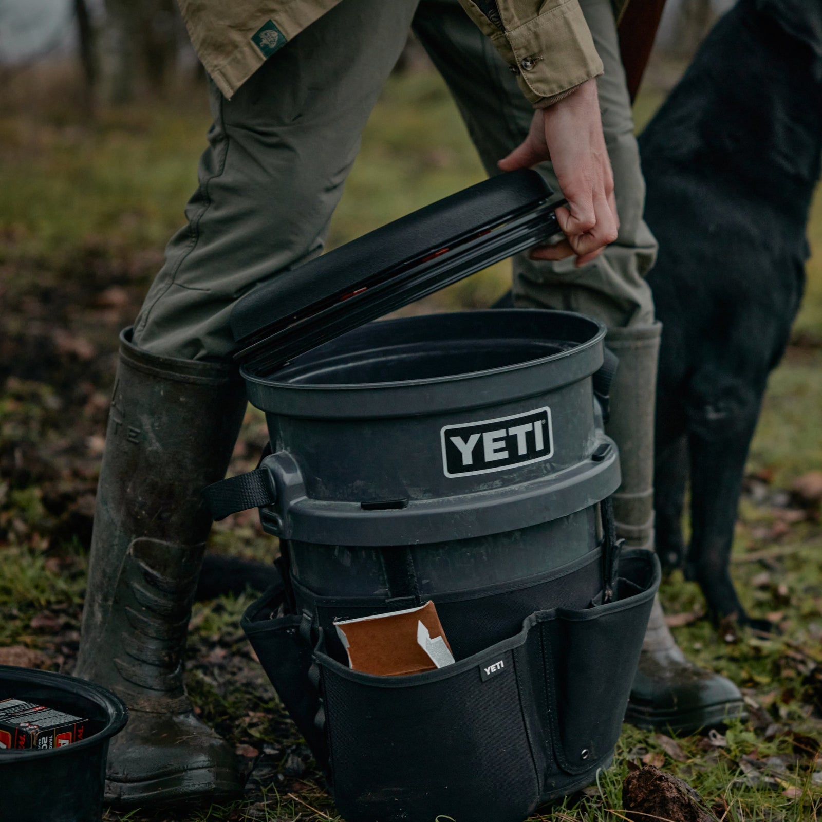 Eine Person in grünen Hosen und Stiefeln öffnet im Freien einen schwarzen YETI® drehbaren Eimerhocker. Der Eimer von YETI ist mit einem Deckel und Beuteln ausgestattet. In der Nähe steht ein schwarzer Hund auf Gras und Erde.