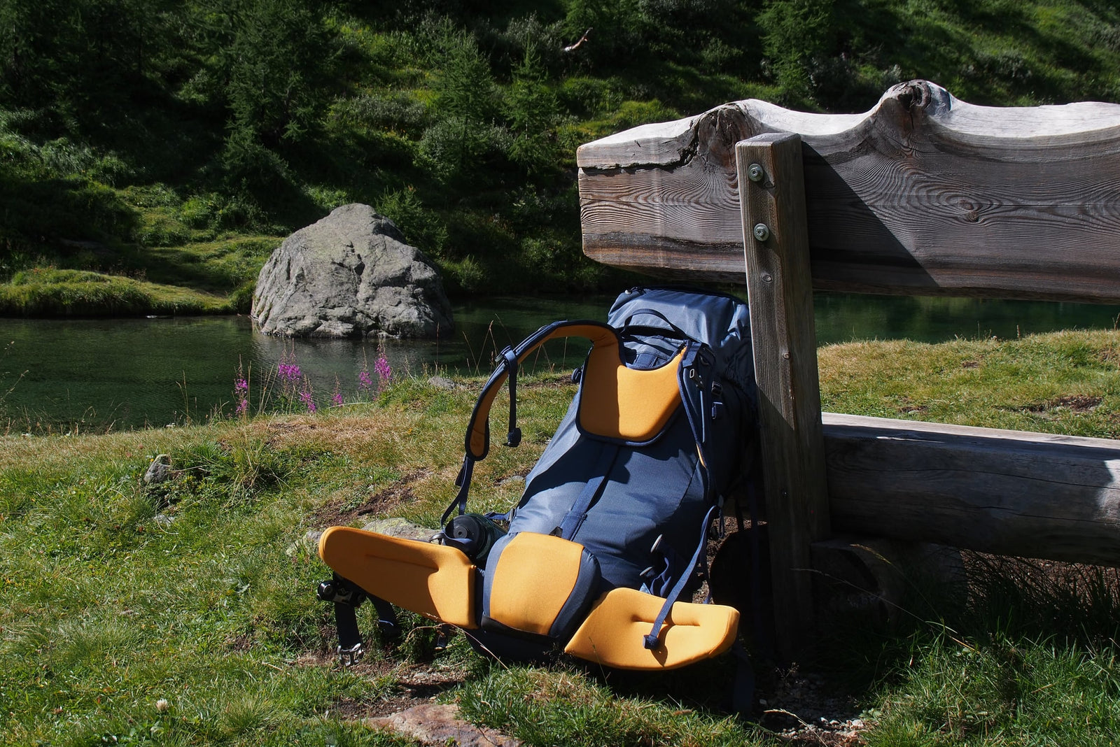 Der blau-orangefarbene EXPED Rucksack Thunder 50 steht neben einer rustikalen Holzbank an einem friedlichen Fluss, umgeben von grünen Bäumen und einem großen Felsen - eine perfekte Wahl von EXPED für Mehrtagestouren.