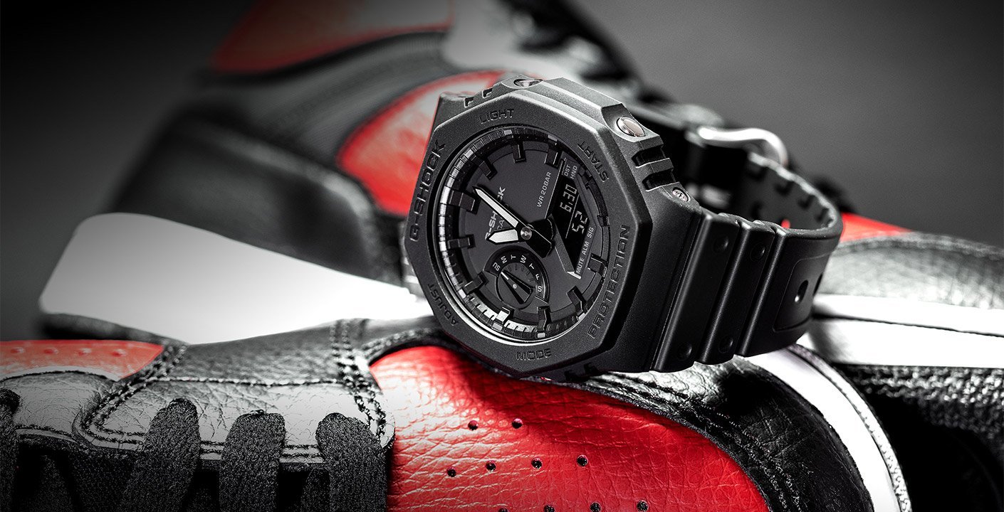 Die G-SHOCK Uhr von G-SHOCK mit ihrem kühnen achteckigen Gehäuse sitzt auf roten und schwarzen Turnschuhen und unterstreicht ihr robustes Design vor einem dramatischen Hintergrund.