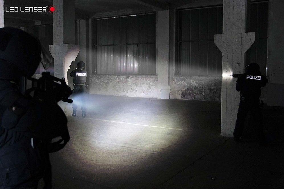 Deutsche Polizeibeamte in taktischer Ausrüstung durchsuchen nachts eine dunkle Lagerhalle und leuchten den Bereich mit LEDLENSER Ledlenser Taschenlampe TT aus, die sie an ihren Waffen befestigt haben.