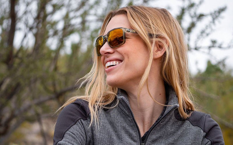 Eine Frau mit langen blonden Haaren trägt eine WILEY X Sonnenbrille ALFA mit abnehmbarem Seitenschutz und eine graue Jacke mit Reißverschluss, während sie lächelnd zur Seite ins Freie blickt, mit verschwommenen Bäumen im Hintergrund.
