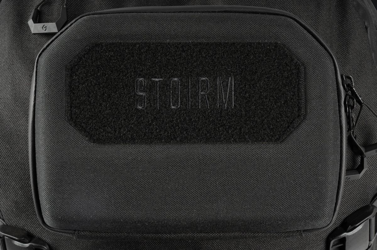 Nahaufnahme des STOIRM 40L Tactical Pack GEN2 in Schwarz mit einem rechteckigen Klettaufnäher mit aufgesticktem "STOIRM" in der Mitte. Dieser robuste, strukturierte Rucksack mit seitlichen Reißverschlüssen ist ideal für raue Outdoor-Abenteuer.