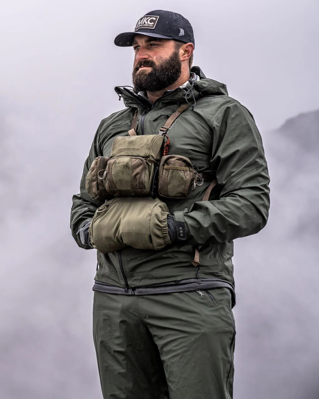Bärtiger Mann in olivgrüner Regenkleidung und MKC-Mütze, der ein taktisches Brustgeschirr mit einem Recon Modular Bino Harness und dem EBERLESTOCK Recon Handwarmer trägt, steht vor einer nebligen Bergkulisse.