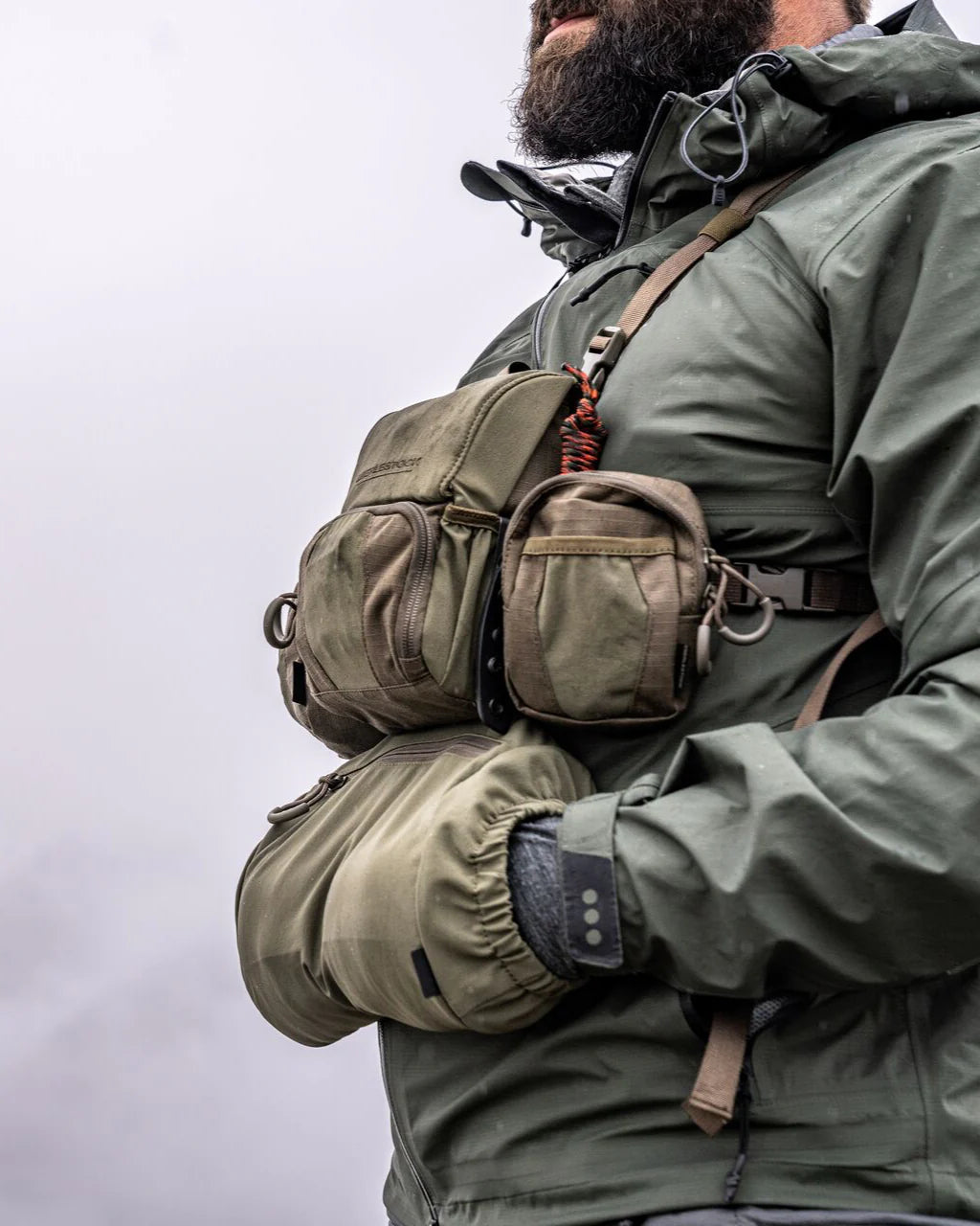 Eine bärtige Person in grüner Outdoor-Jacke steht vor einem wolkenverhangenen Himmel. Sie trägt isolierte Handschuhe und den EBERLESTOCK Recon Handwarmer, der an einem modularen Bino-Gurt befestigt ist.