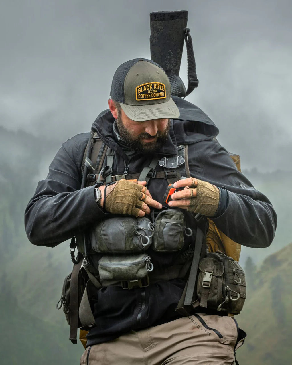 Ein bärtiger Mann mit einer Mütze der Black Rifle Coffee Company und Handschuhen passt seine taktische Weste im Freien an und befestigt eine EBERLESTOCK Recon Utility Pouch daran. Er trägt ein Gewehr auf dem Rücken und steht in einer nebligen Berglandschaft.