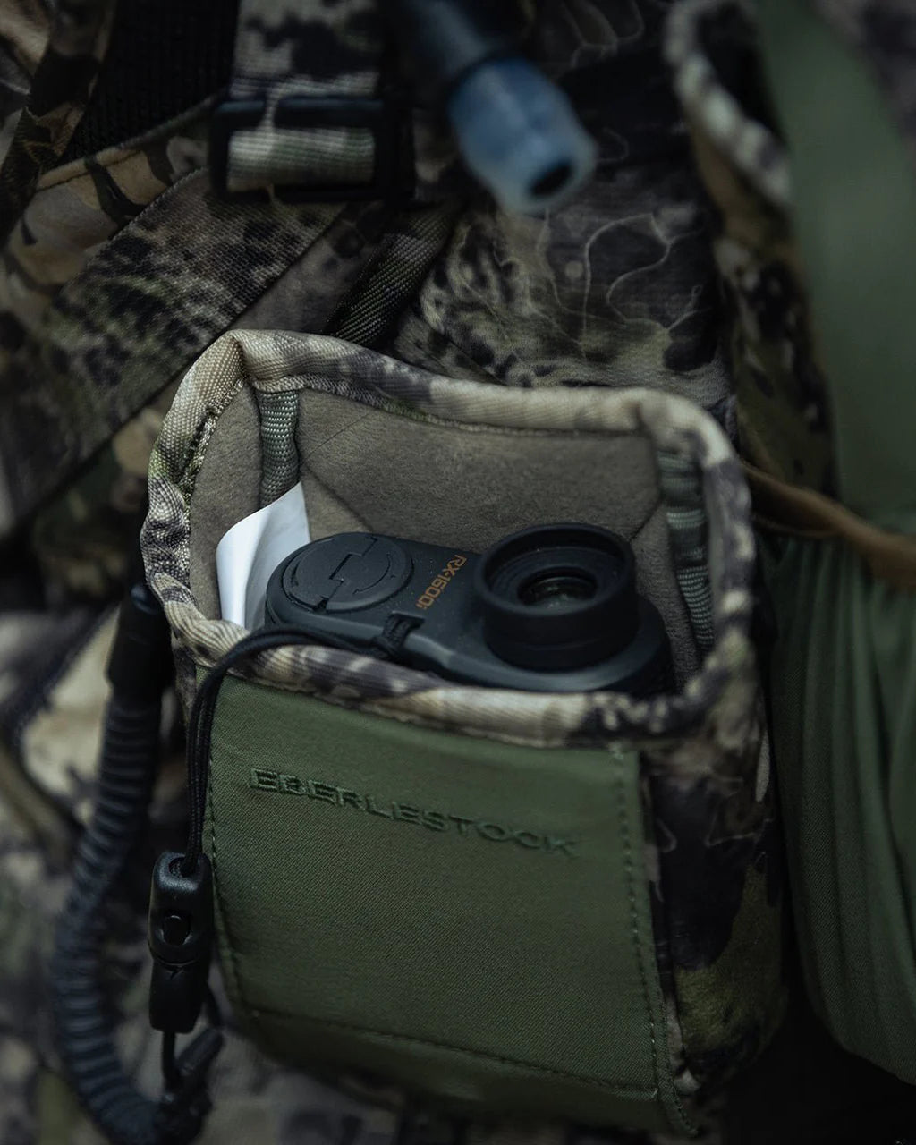Die EBERLESTOCK Recon Rangefinder Pouch in Camouflage fasst einen kompakten schwarzen Entfernungsmesser und einen kleinen weißen Gegenstand, der sicher an der Outdoor-Ausrüstung oder dem Recon Modular Bino Harness mit passendem Tarnmuster befestigt ist.