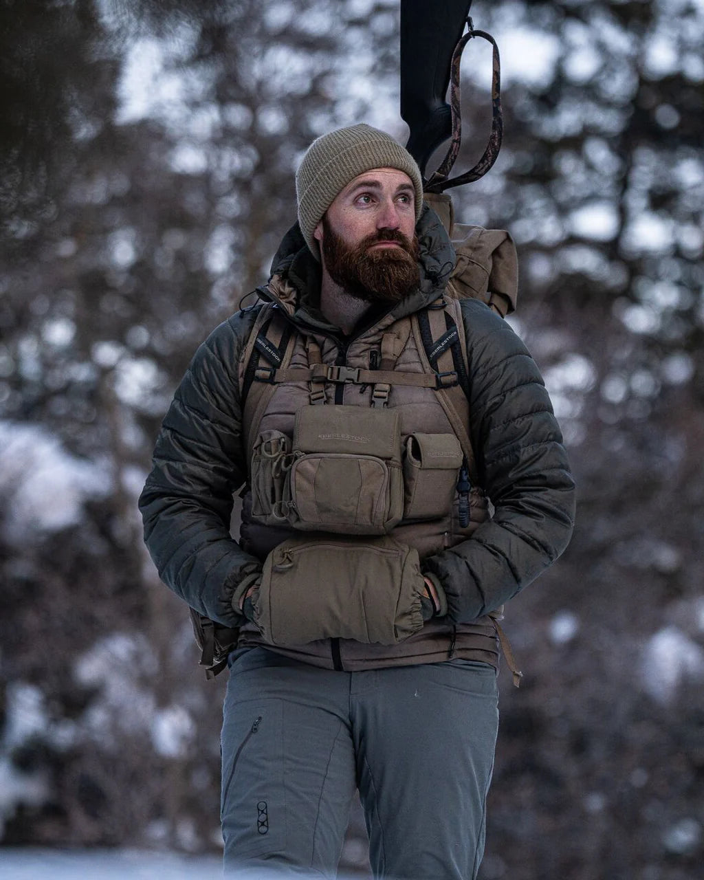 Ein bärtiger Mann in Outdoor-Ausrüstung steht in einem verschneiten Wald mit dem EBERLESTOCK Recon Handwarmer auf der Brust, trägt eine beigefarbene Mütze und eine wattierte Jacke und schaut zur Seite.