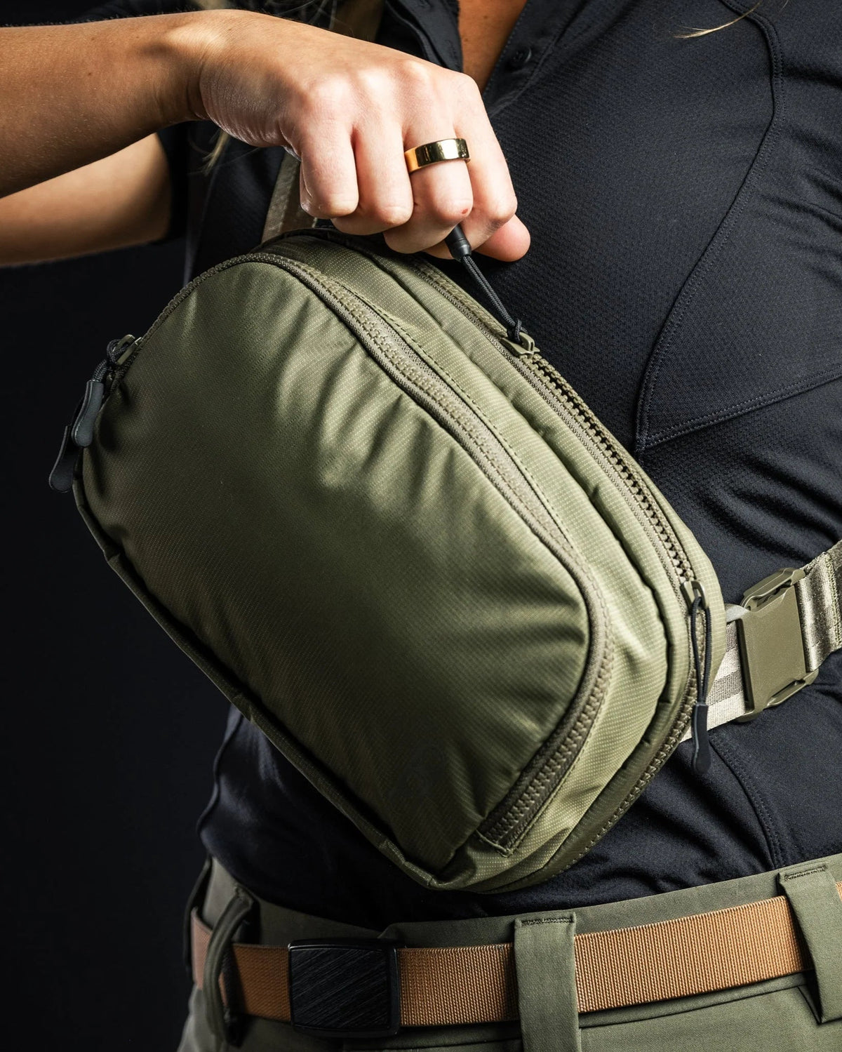 Eine Person mit einem schwarzen Hemd und einem goldenen Ring öffnet den Reißverschluss einer olivgrünen EBERLESTOCK Fade Flex Crossbody Bag. Die kompakte Tasche hat mehrere Fächer mit Reißverschluss und einen verstellbaren Schultergurt.