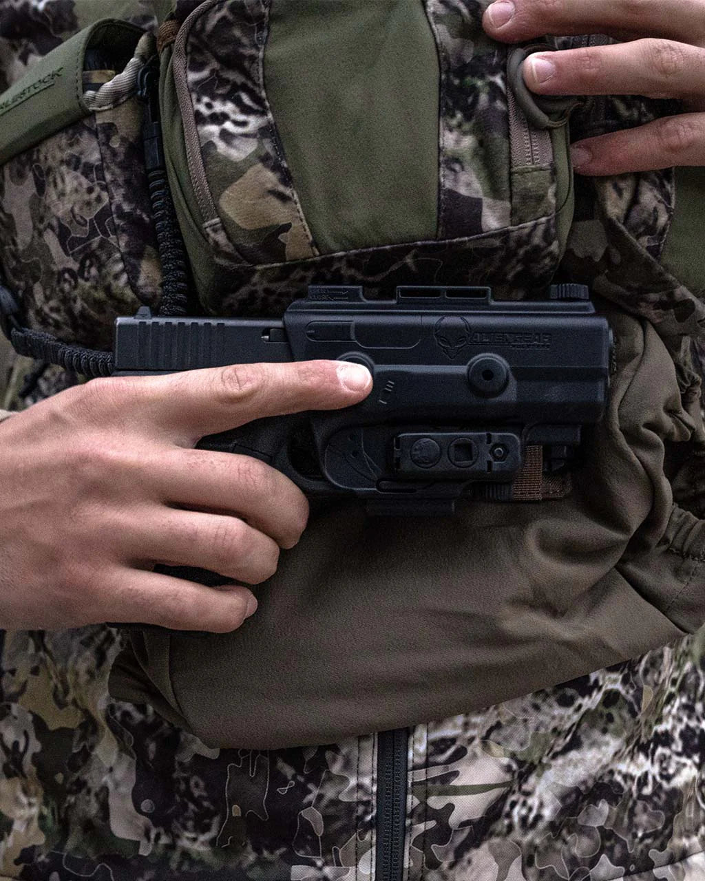 Eine Person in Camouflage hält eine schwarze Handfeuerwaffe in einem Holster mit einem Finger am Abzugsbügel, während sie ein EBERLESTOCK Recon MOLLE Panel auf ihrer Brust einstellt.