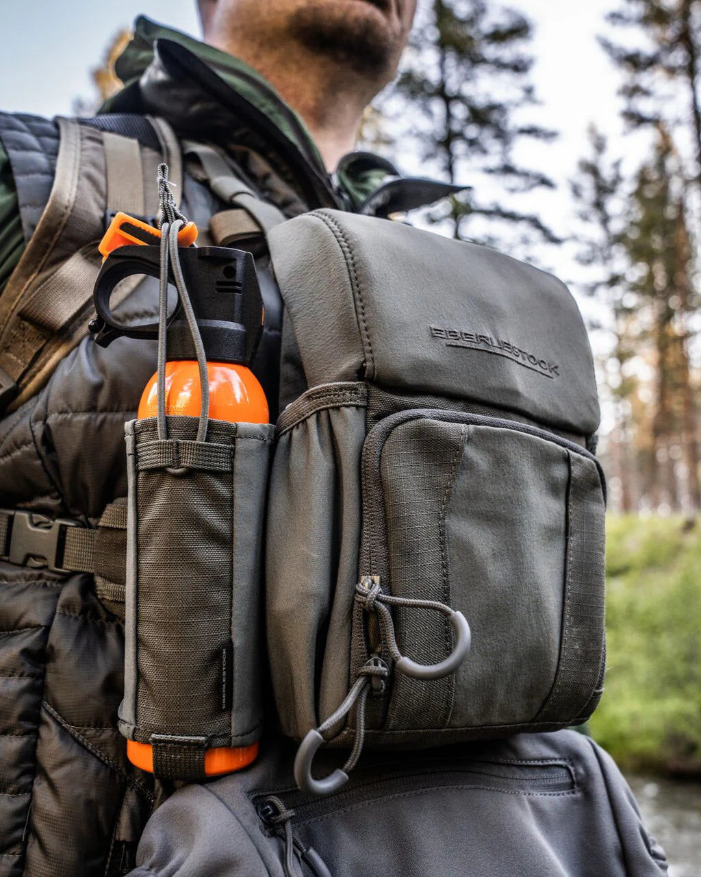 Nahaufnahme einer Person im Freien mit einem EBERLESTOCK-Rucksack. Eine leuchtend orangefarbene Wasserflasche und ein Eberlestock Recon Bear Spray Pouch sind in einer Seitentasche gesichert, umgeben von Bäumen und sanftem Sonnenlicht.