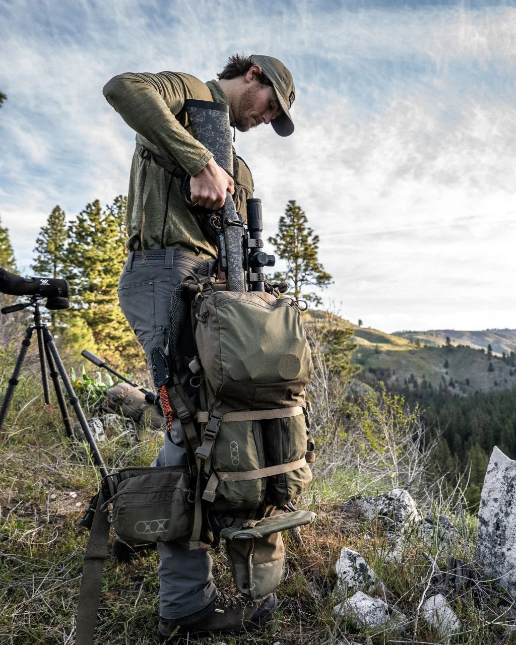 Ein Mann in Outdoor-Ausrüstung steht an einem felsigen Berghang und packt ein Gewehr in einen EBERLESTOCK Batwing V2 Rucksack. In der Nähe befinden sich ein Stativ und ein Spektiv, mit bewaldeten Hügeln und einem bewölkten Himmel im Hintergrund.